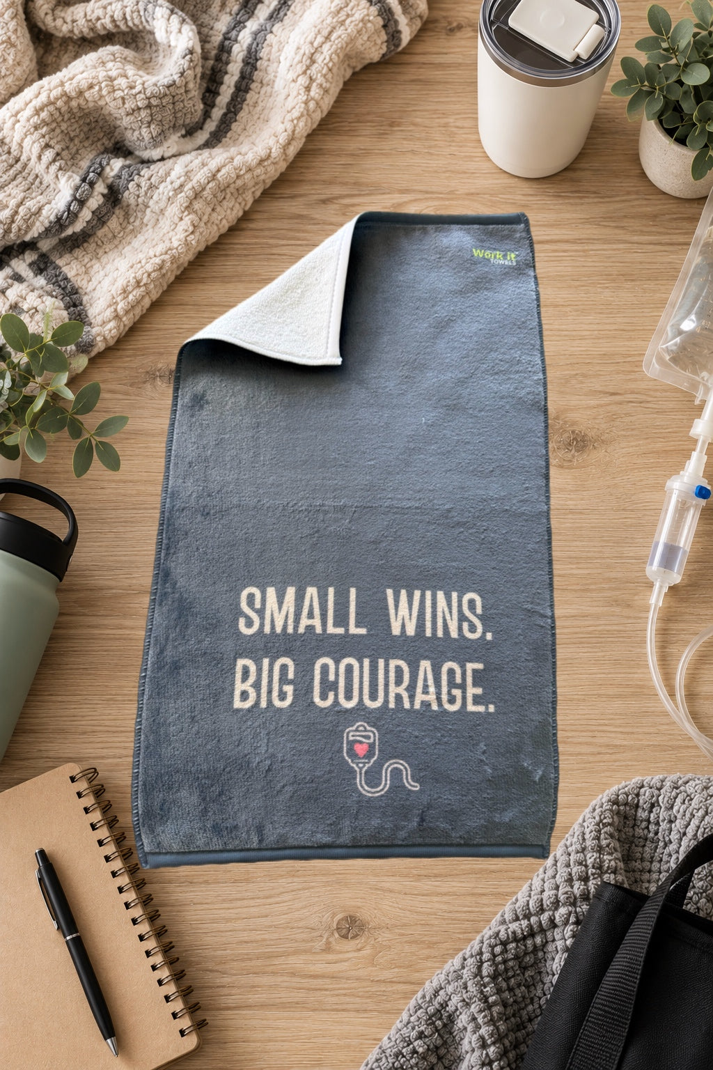 blueish grey plush towel with text, 'small wins, big courage,' and a small IV graphic with a heart on a hardwood floor with blankets and drinks