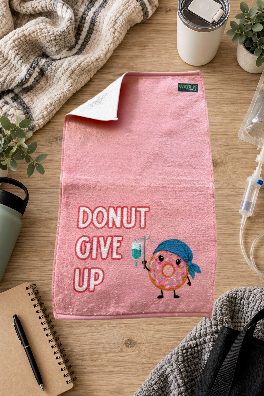 plush pink towel with 'donut give up' and a pink iced donut wearing a cap and holding an IV on a hardwood floor surrounded by blankets, a notebook and drinks.