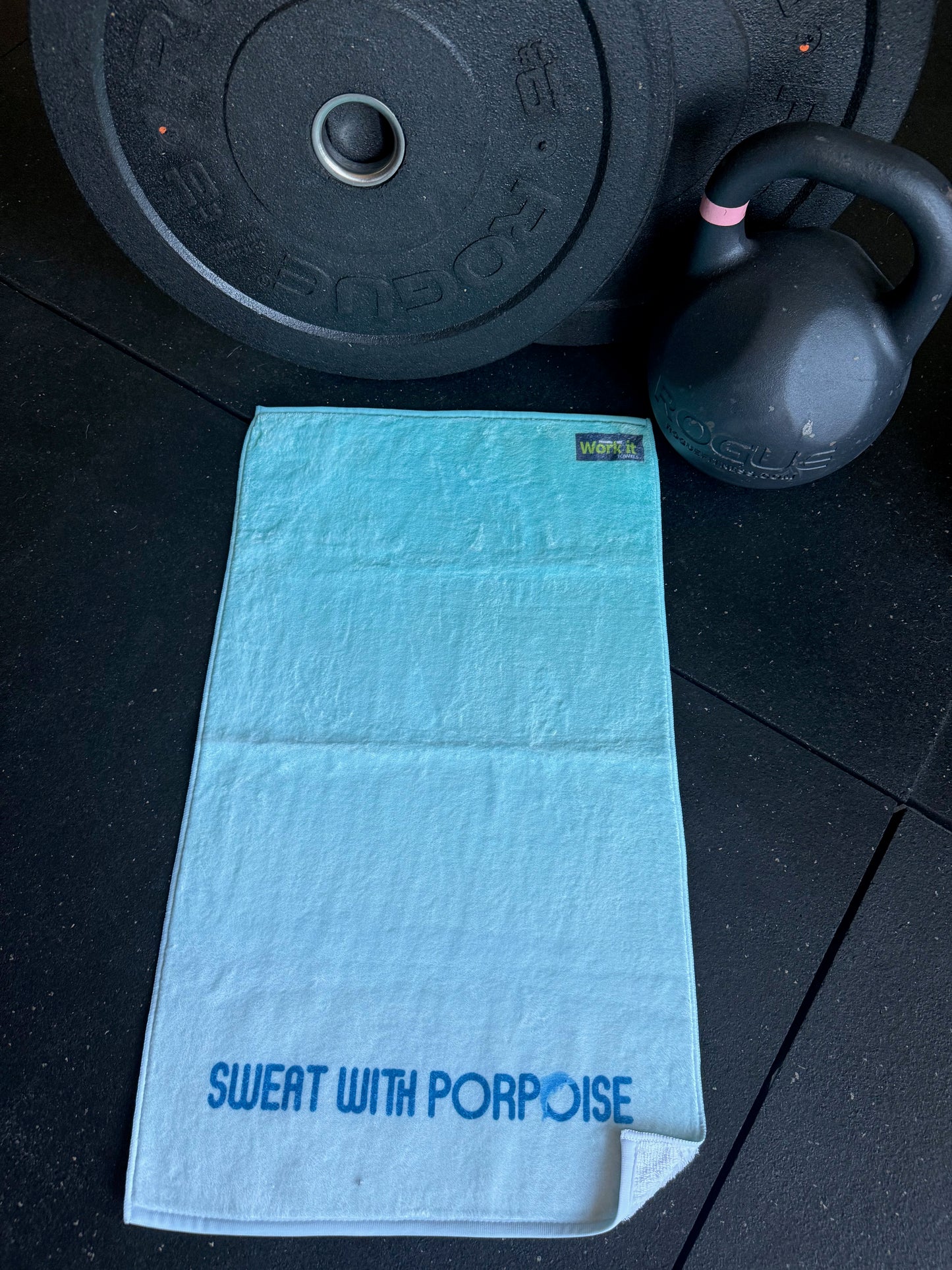 Blue towel with 'Sweat with Porpoise' text on a black floor with weights in the background