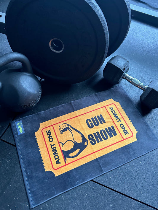 A black gym floor with a kettlebell, barbell plates, and a dumbbell surrounding a black workout towel printed with an orange “Admit One Gun Show” ticket design featuring a flexed bicep illustration.