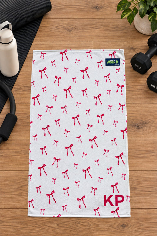 Pink bow personalized gym towel with custom 'KP' initials, laid flat on a wood floor next to fitness gear.