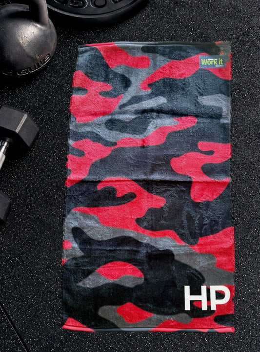 Red camouflage personalized gym towel with custom 'HP' initials, laid flat on a black gym floor with a kettlebell and dumbbell.