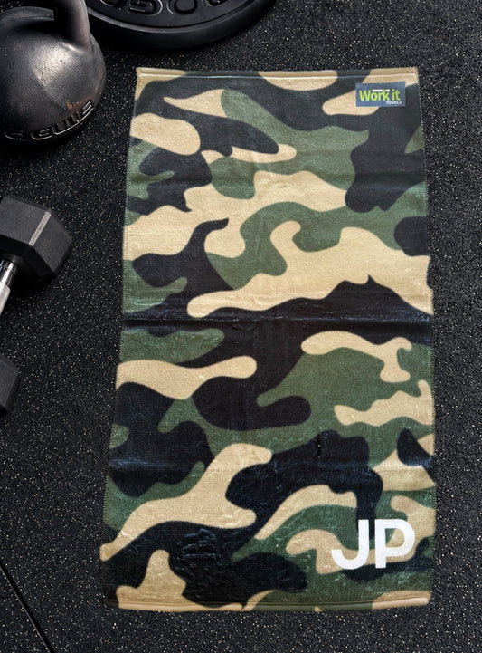 Green camouflage personalized gym towel with custom 'JP' initials, laid flat on a black gym floor with a kettlebell and dumbbell.