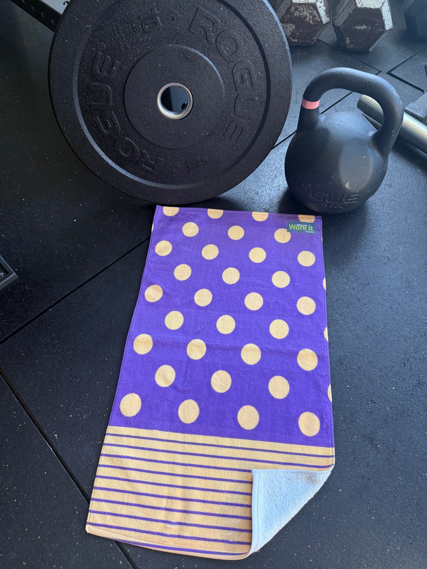 Purple towel with gold polka dots and stripes on a gym floor with weights and a kettlebell.