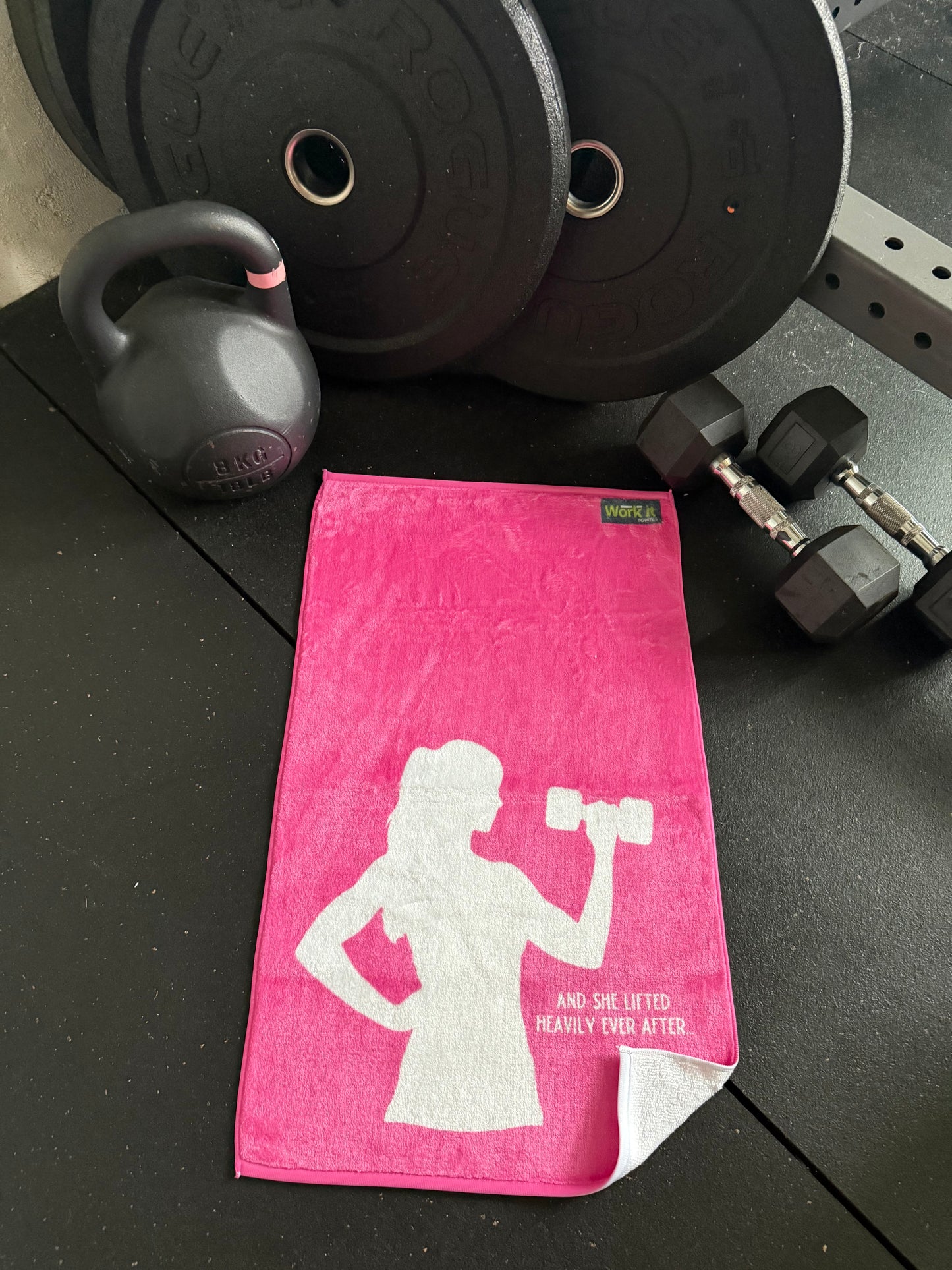 Pink towel with white woman graphic and heavily ever after on a black surface with gym equipment
