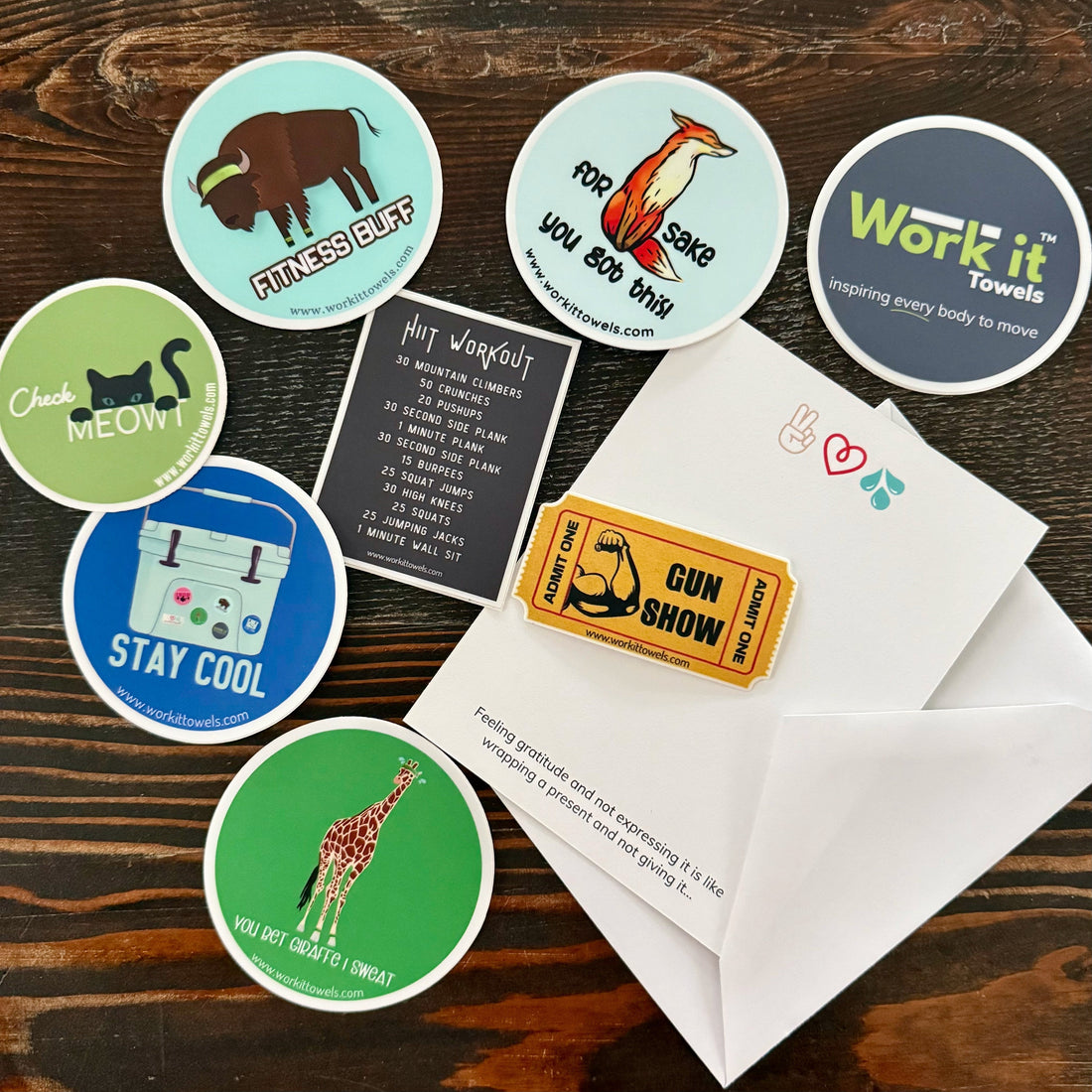 various bright, punny stickers on a wooden coffee table displayed with a thank you note.  Featuring punny Work It Towels designs.