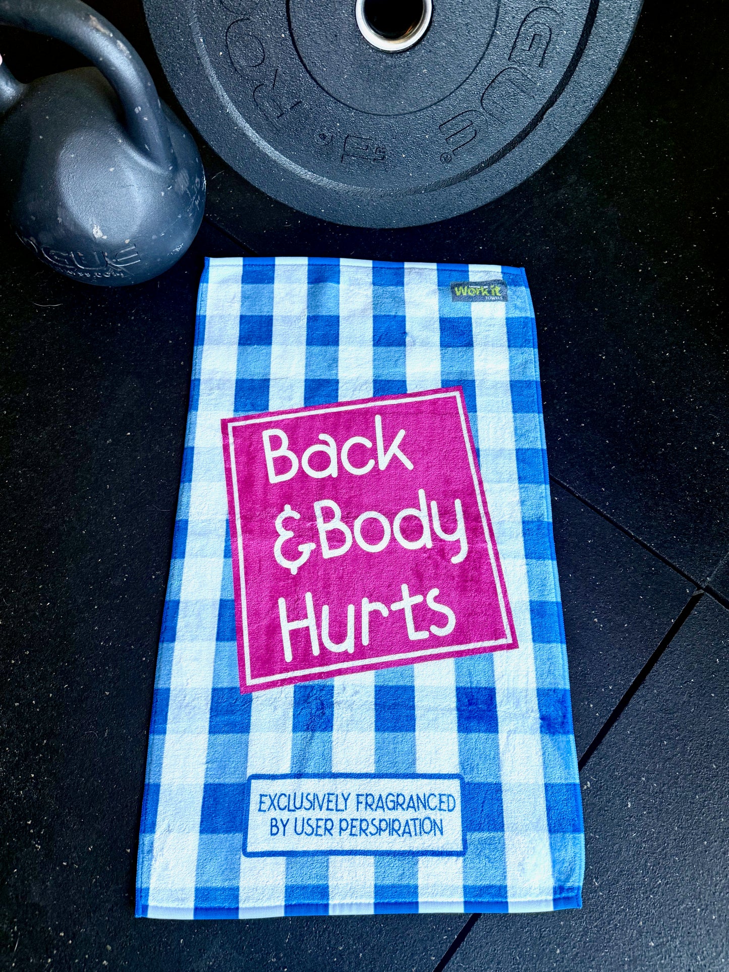 Blue and white checkered towel with 'Back & Body Hurts' text on a dark surface with gym equipment.