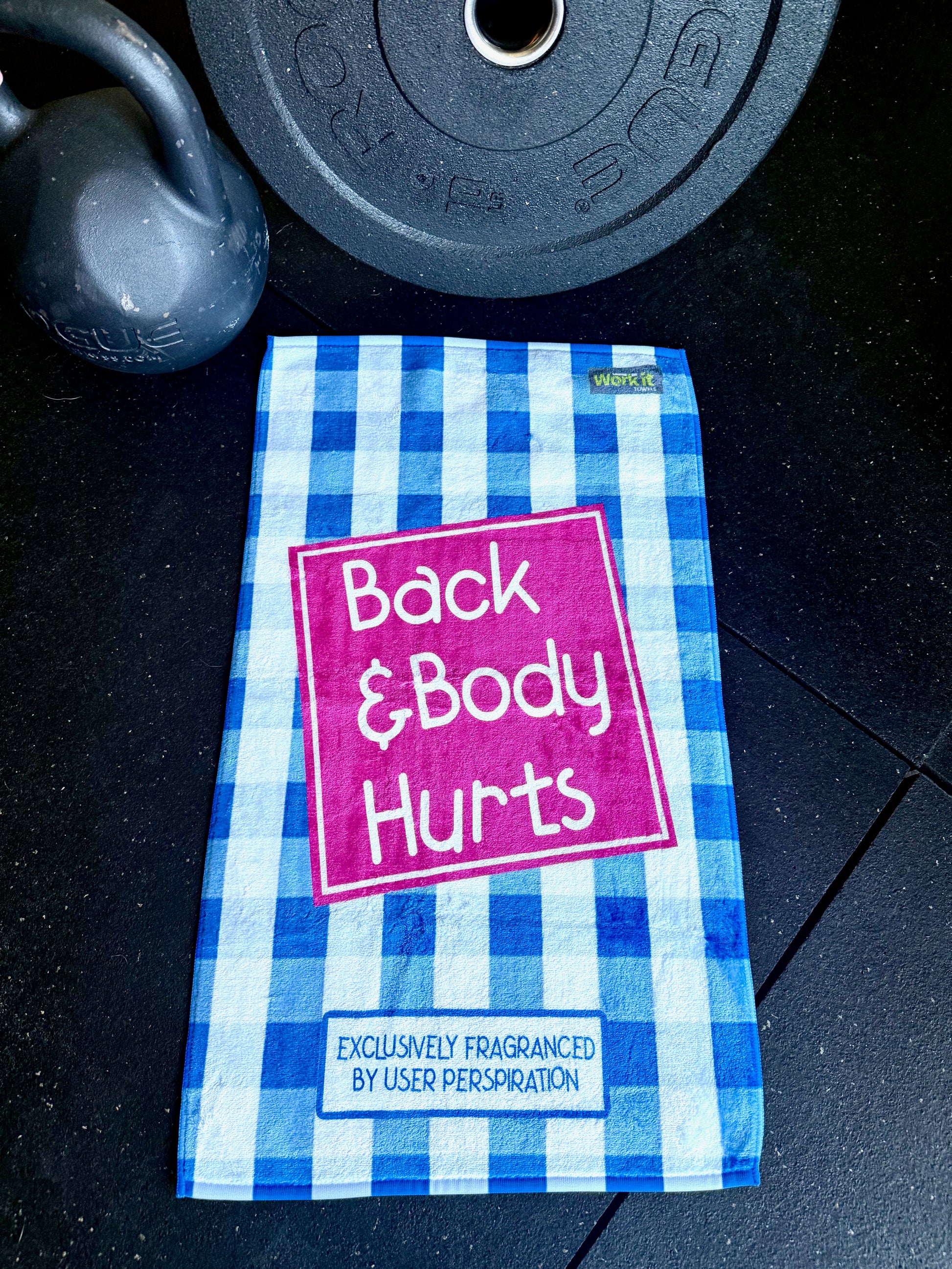 Blue and white checkered towel with 'Back & Body Hurts' text on a dark surface with gym equipment.