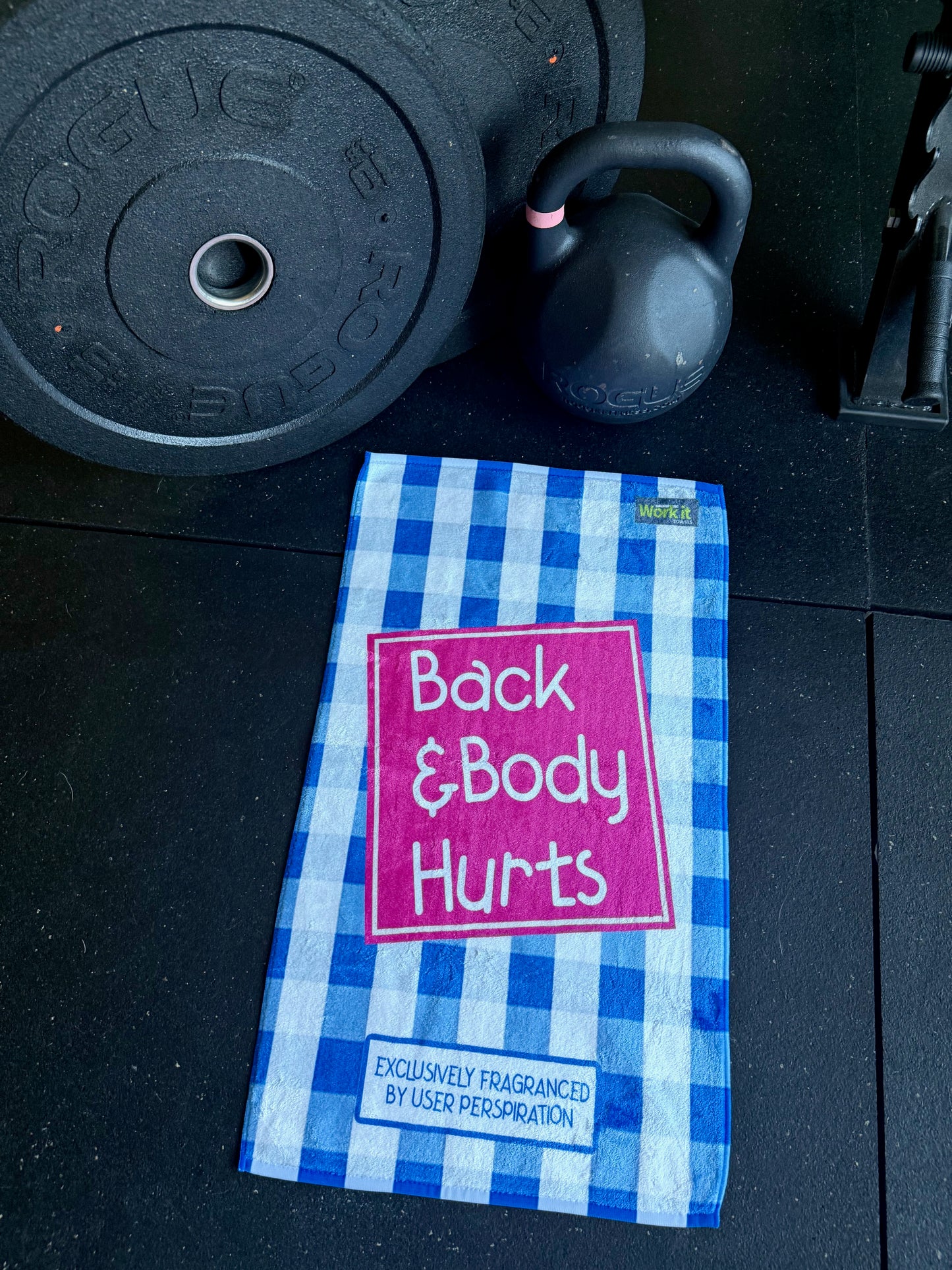Blue and white checkered towel with 'Back & Body Hurts' text on a gym floor with weights.