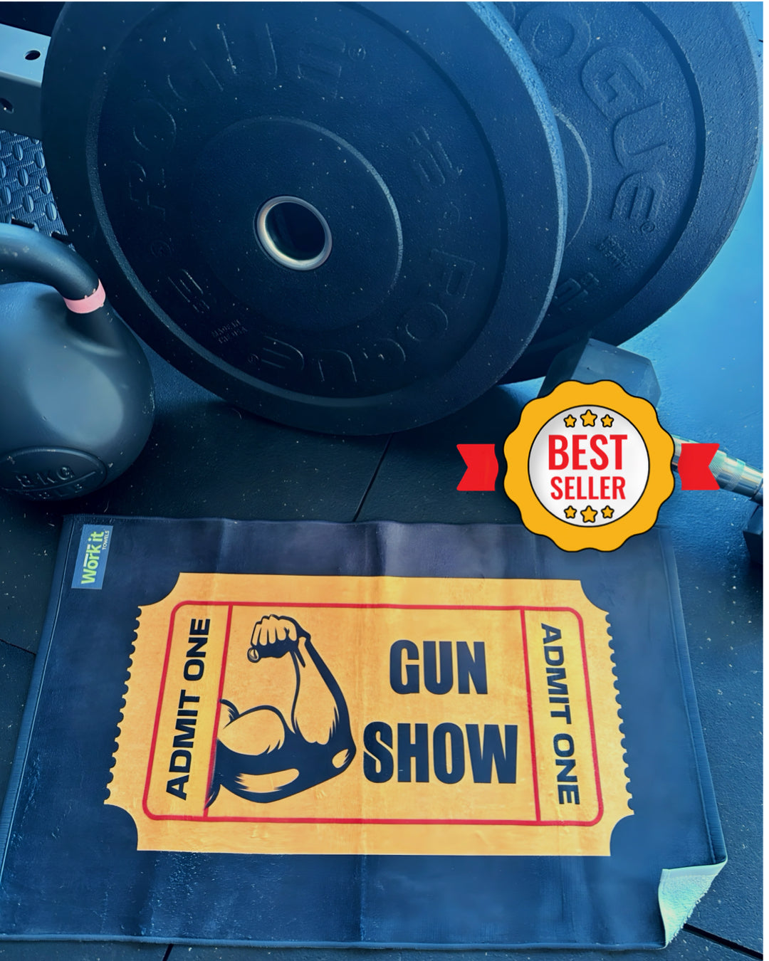 Black gym mat with an image of a gym towel with a bicep flexing and 'admit one' yellow ticket design to the 'Gun Show' with a 'Best Seller' badge, next to gym equipment.