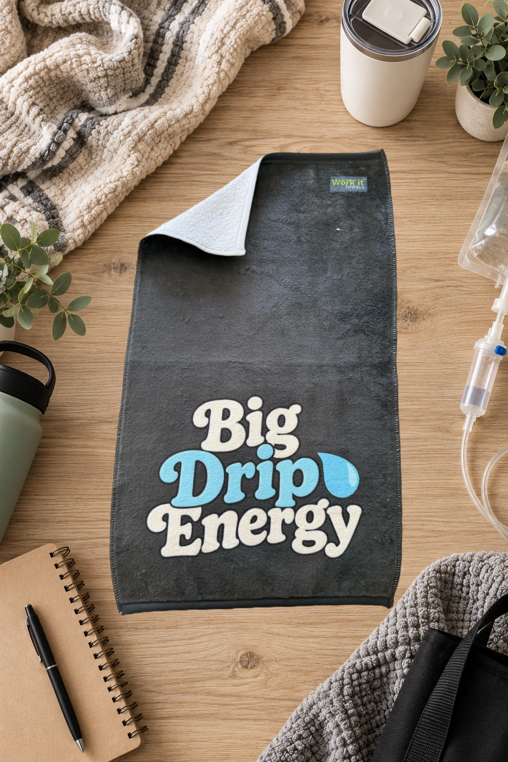 dark grey towel with 'big drip energy' text and a tear drop on a hardwood floor with blankets and drinks