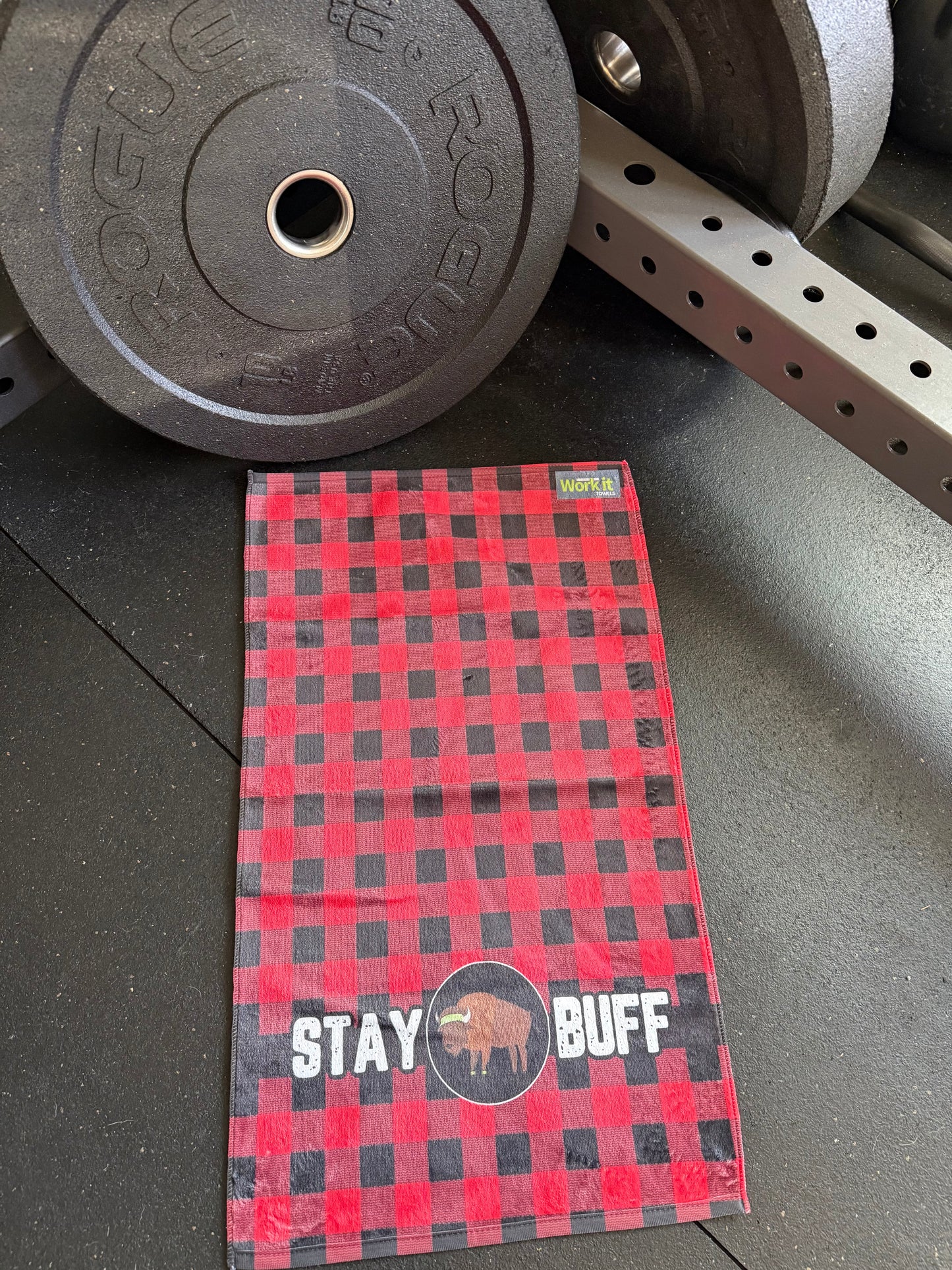 Red and black checkered towel with 'Stay Buff' text on a gym floor with weights in the background