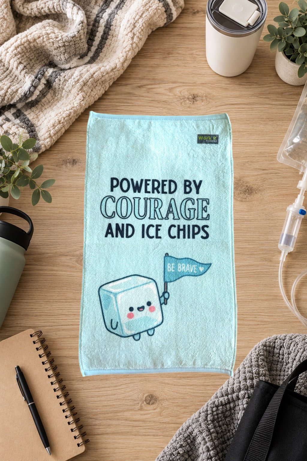 A light blue towel laid flat on a wooden table, printed with the phrase 'POWERED BY COURAGE AND ICE CHIPS' and a cute smiling ice cube character holding a flag that says 'BE BRAVE'. The scene is styled with a knit blanket, plants, a notebook, pen, tumbler, and medical tubing around it.