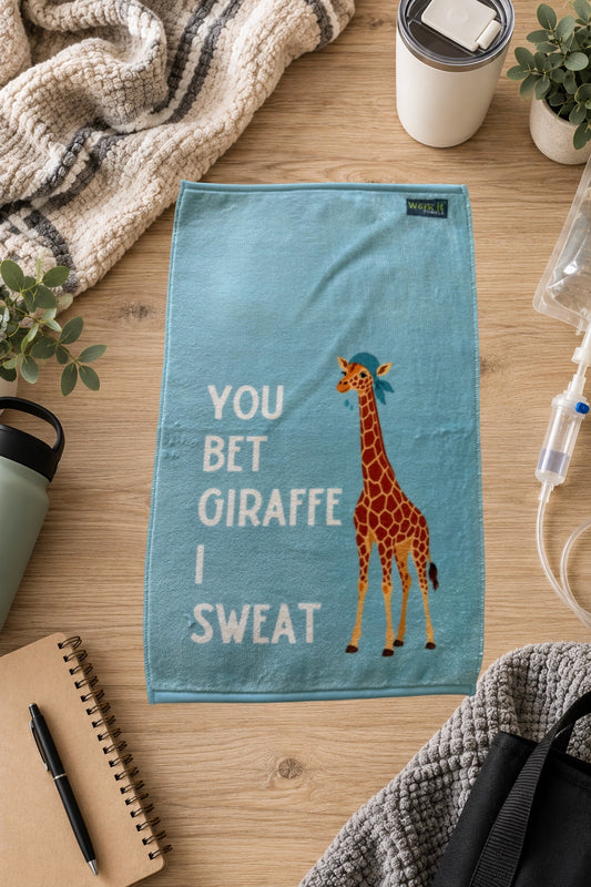 a light blue towel with a giraffe wearing a cap and sweating with text, 'you bet giraffe I sweat,' on a hardwood floor with blankets and drinks. 