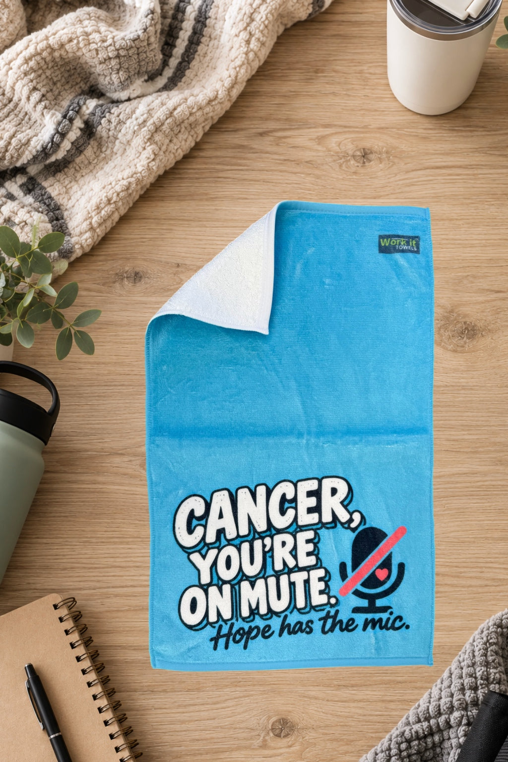 Blue Cancer, You're On Mute towel with one corner folded back to show the soft white terry backing, on a wood table with a blanket and drinkware.