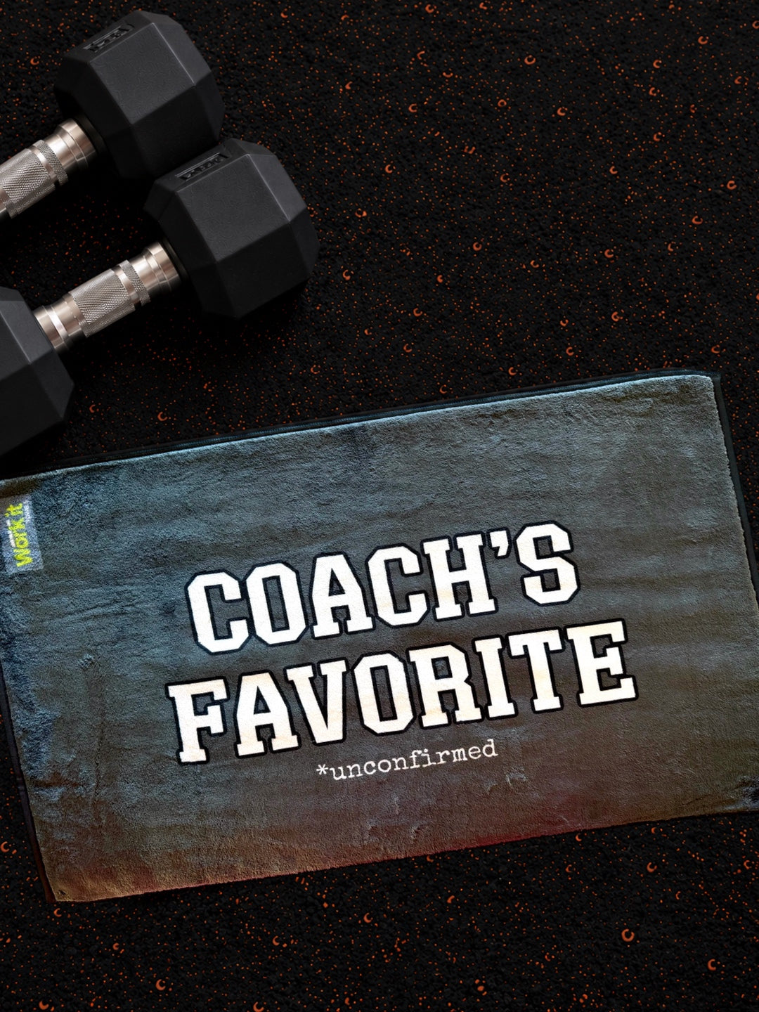plush grey gym towel with 'coach's favorite unconfirmed' in white font on a dark gym floor with hand weights