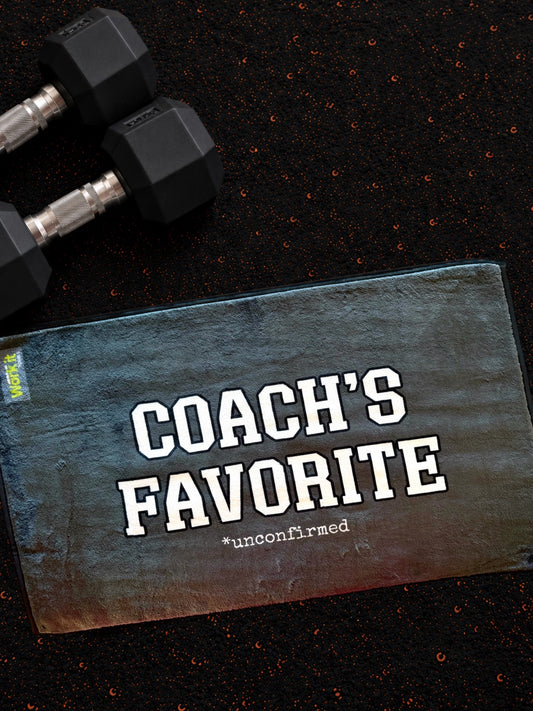 plush grey gym towel with 'coach's favorite unconfirmed' in white font on a dark gym floor with hand weights