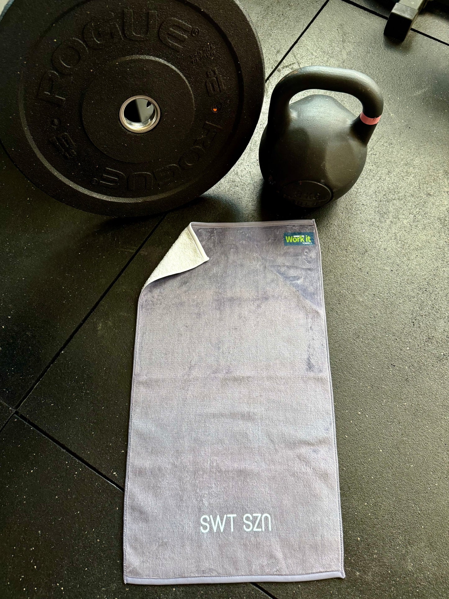 Gray towel with ‘SWT SZN’ text on a floor next to a kettlebell and weight plate