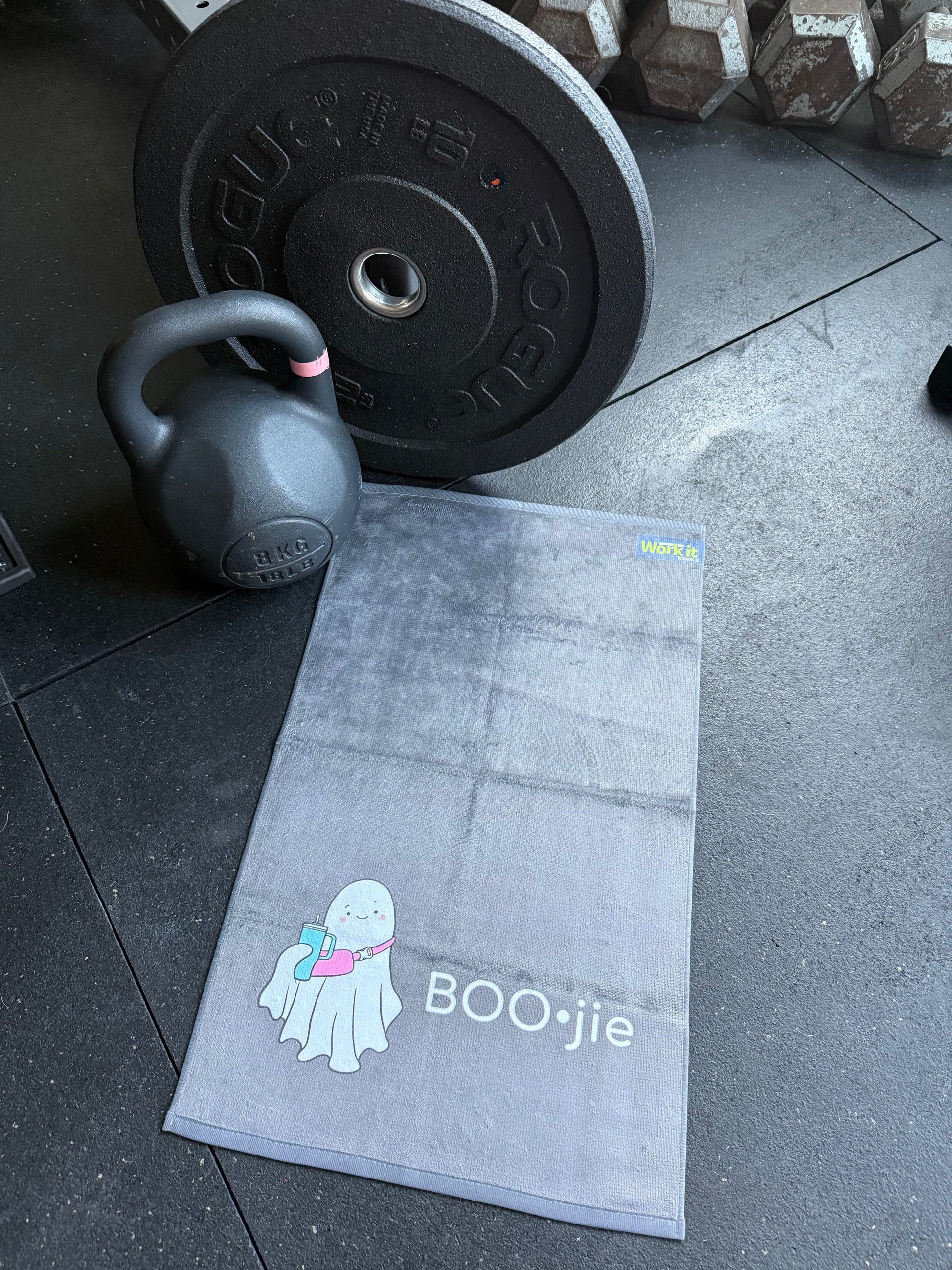 Gray exercise towel with 'BOO•jie' branding on a gym floor with kettlebell and barbell.