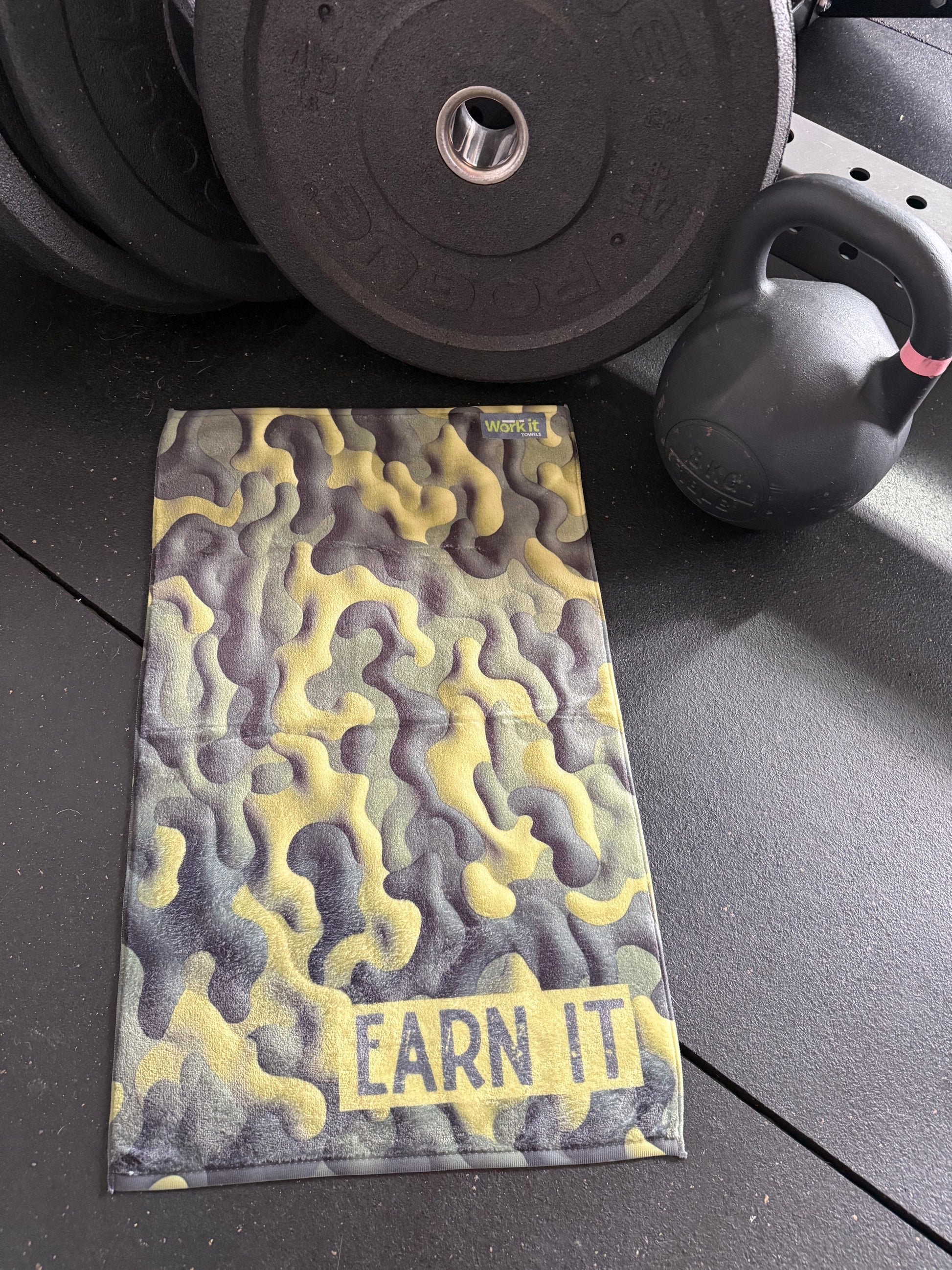 Camouflage towel with 'Earn It' text on a black surface with gym equipment.