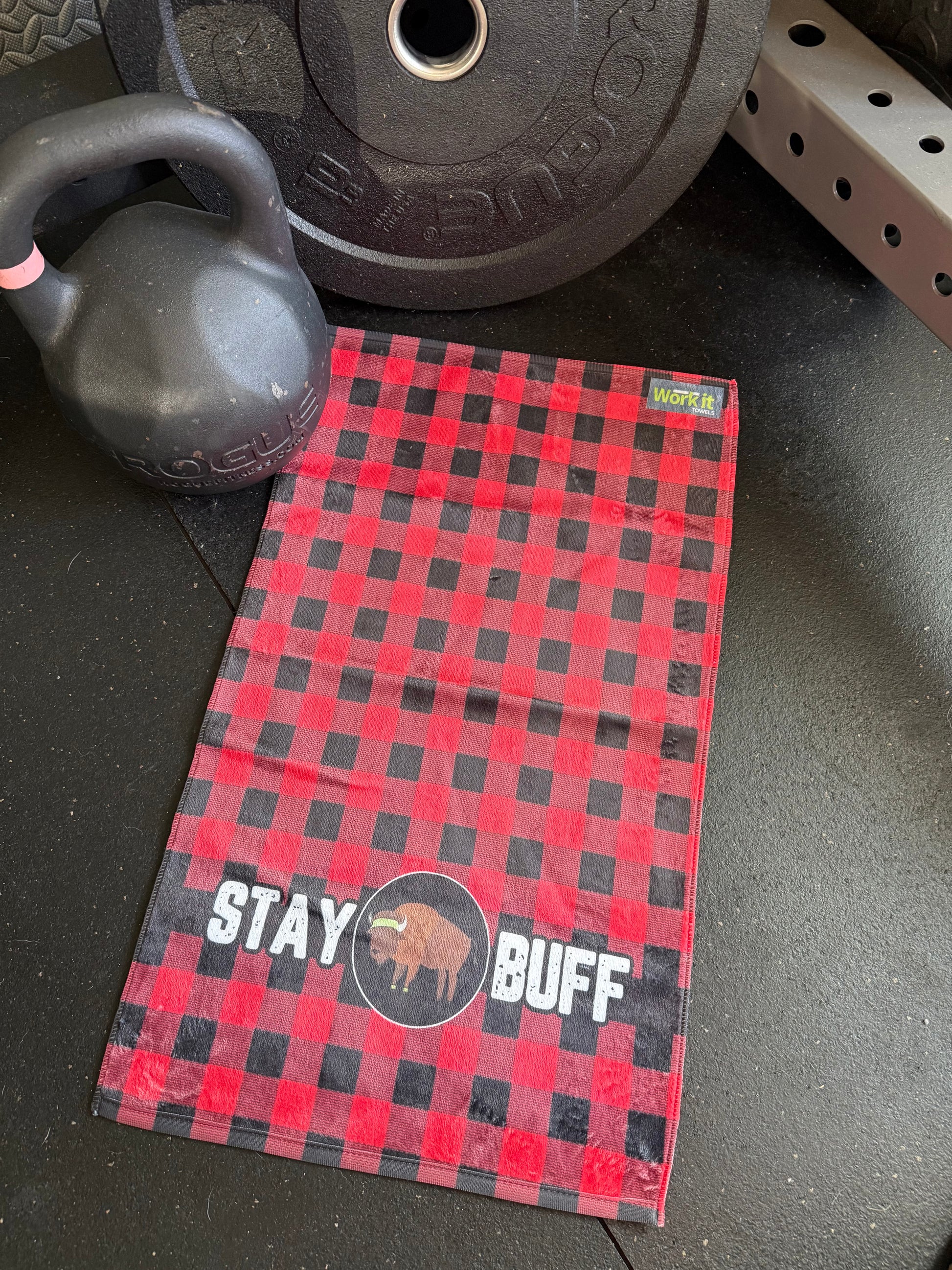 Red and black checkered towel with 'Stay Buff' text on a gym floor with weights and kettlebell.