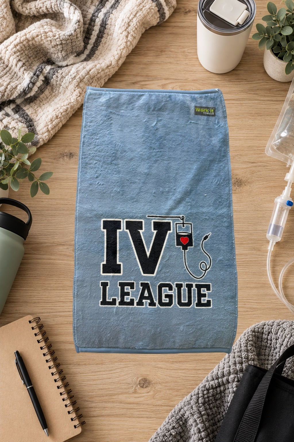 blueish grey towel with text, 'IV League' and an IV hanging from the V.  The towel is laying on a hardwood floor with blankets and IVs