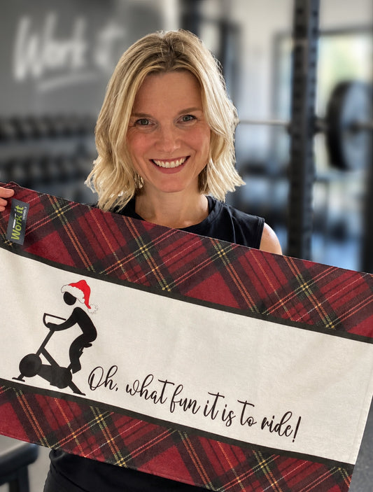 A smiling blonde woman in a gym holds up a red and black plaid scarf with a white center panel featuring a black scooter graphic and the text 'Oh, what fun it is to ride!'.
