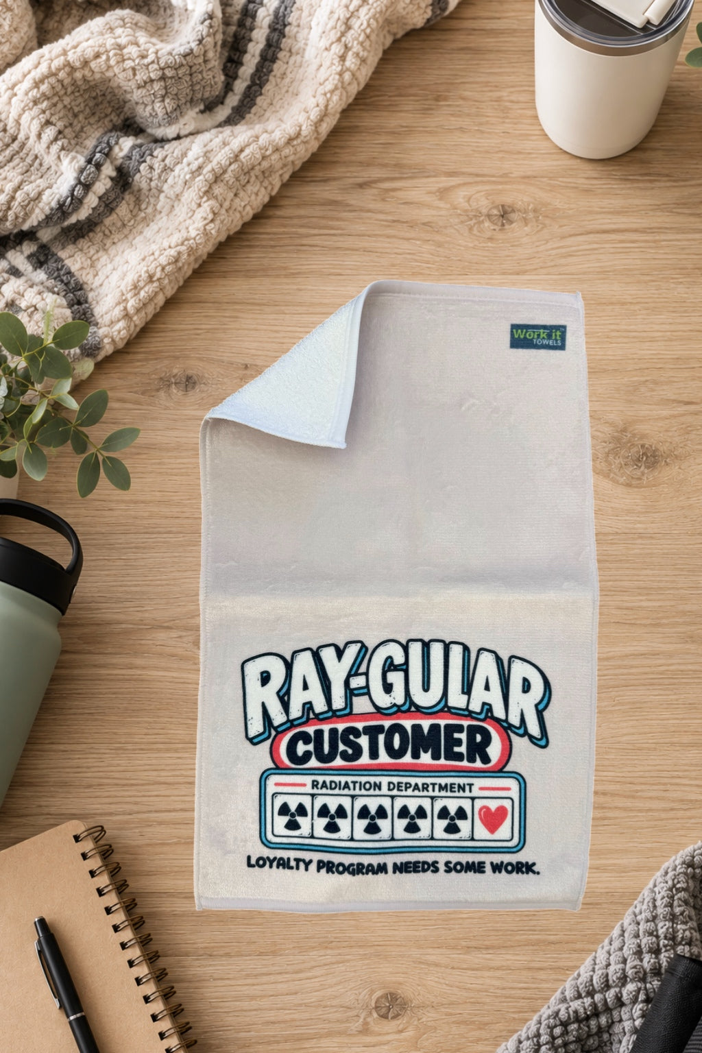 Cream Ray-gular Customer towel with one corner folded back to show the soft white terry backing, on a wood table with a blanket and drinkware.