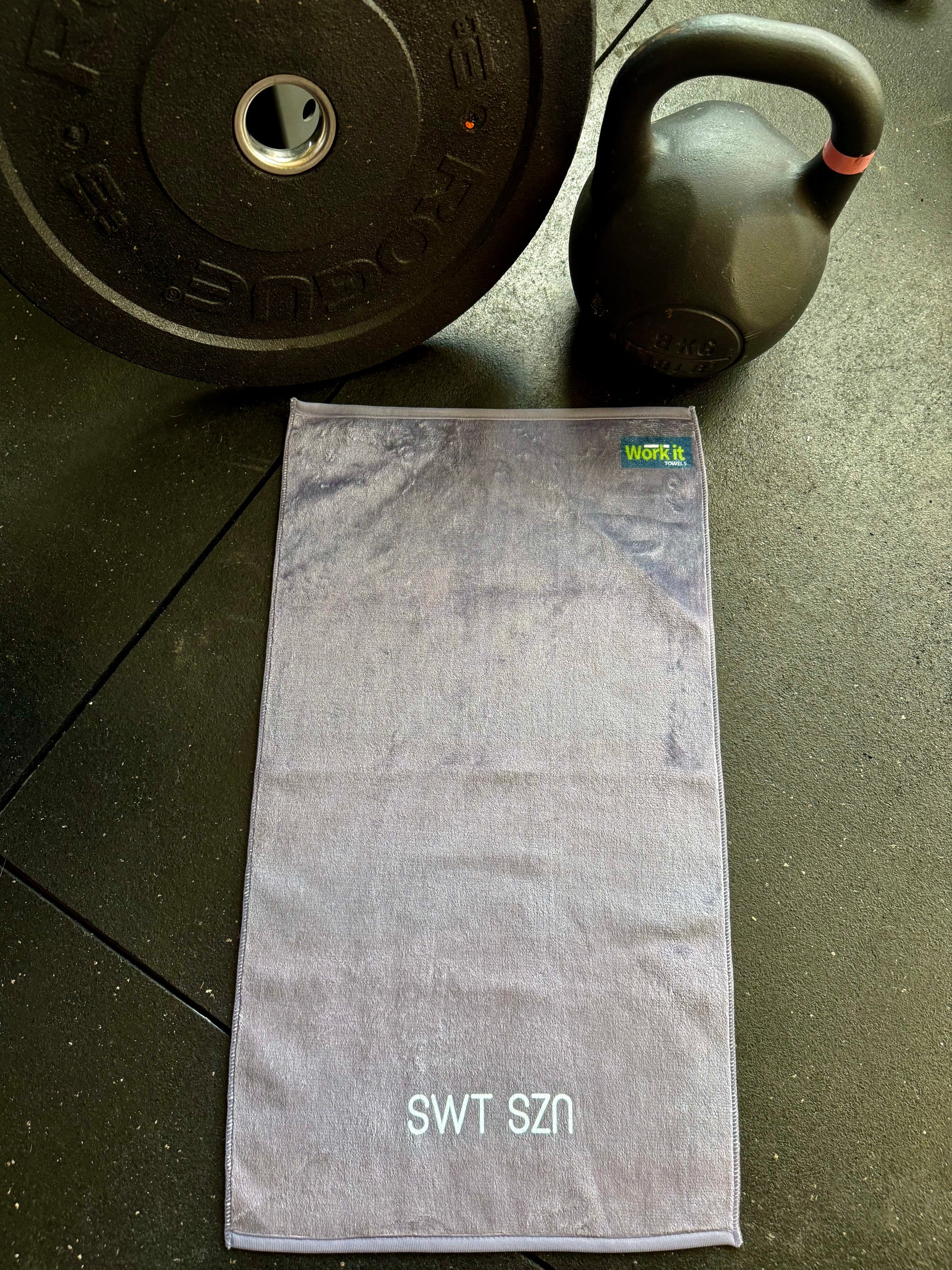 Gray towel with 'SWT SZN' text on a dark surface with a kettlebell and weight plate.