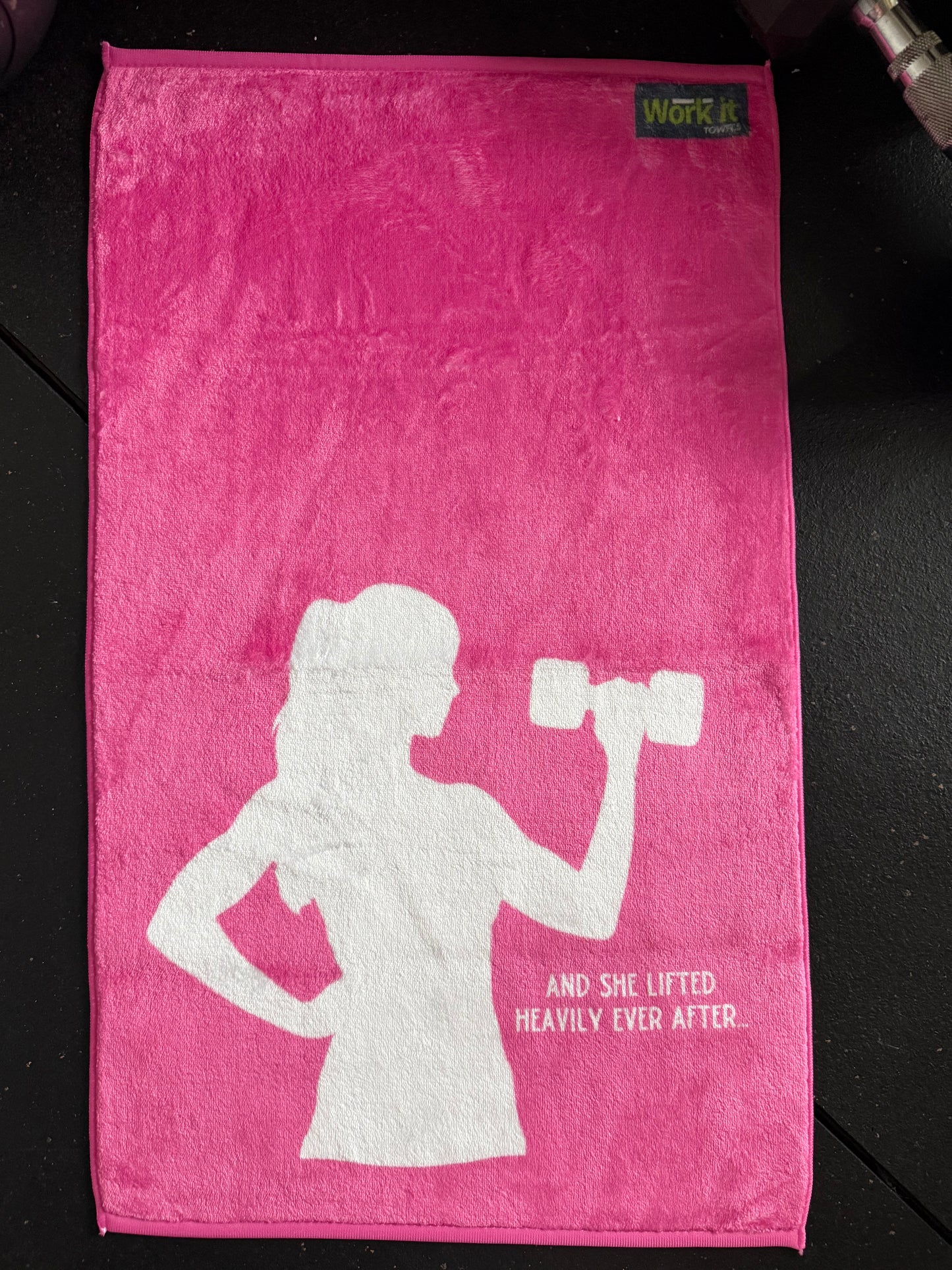 Pink towel with white silhouette of a woman lifting weights and text on a dark surface