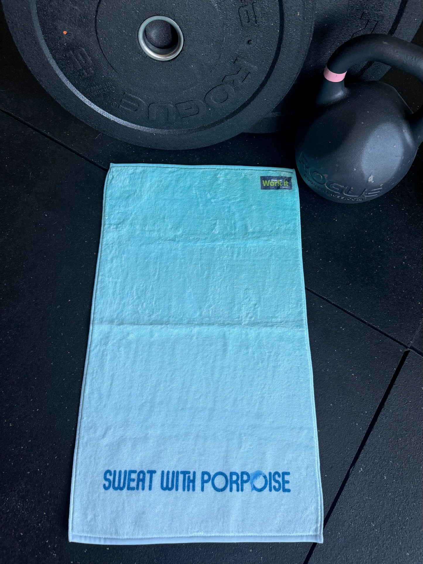 Blue towel with 'Sweat with Porpoise' text on a black floor with gym equipment.
