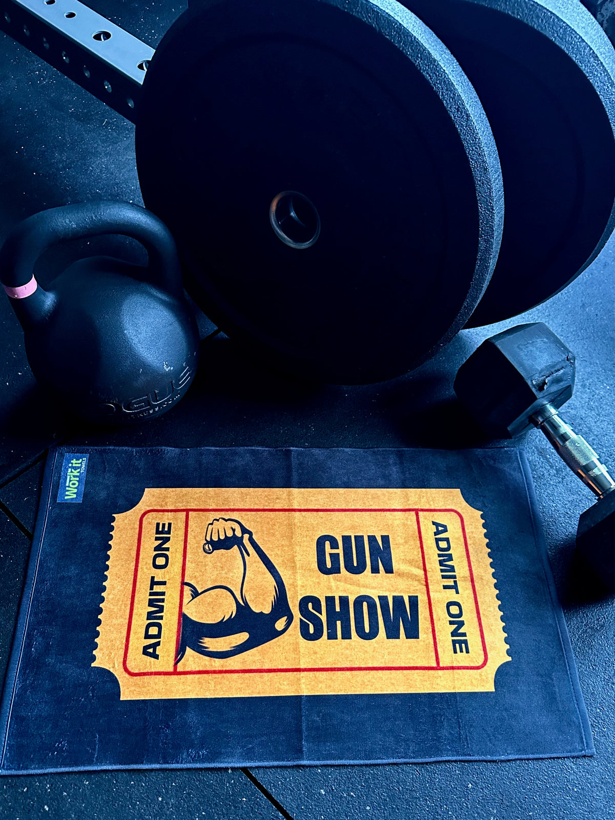 black gym towel with yellow admit one ticket and 'gun show' with flexed bicep design towel on a gym floor with gym accessories