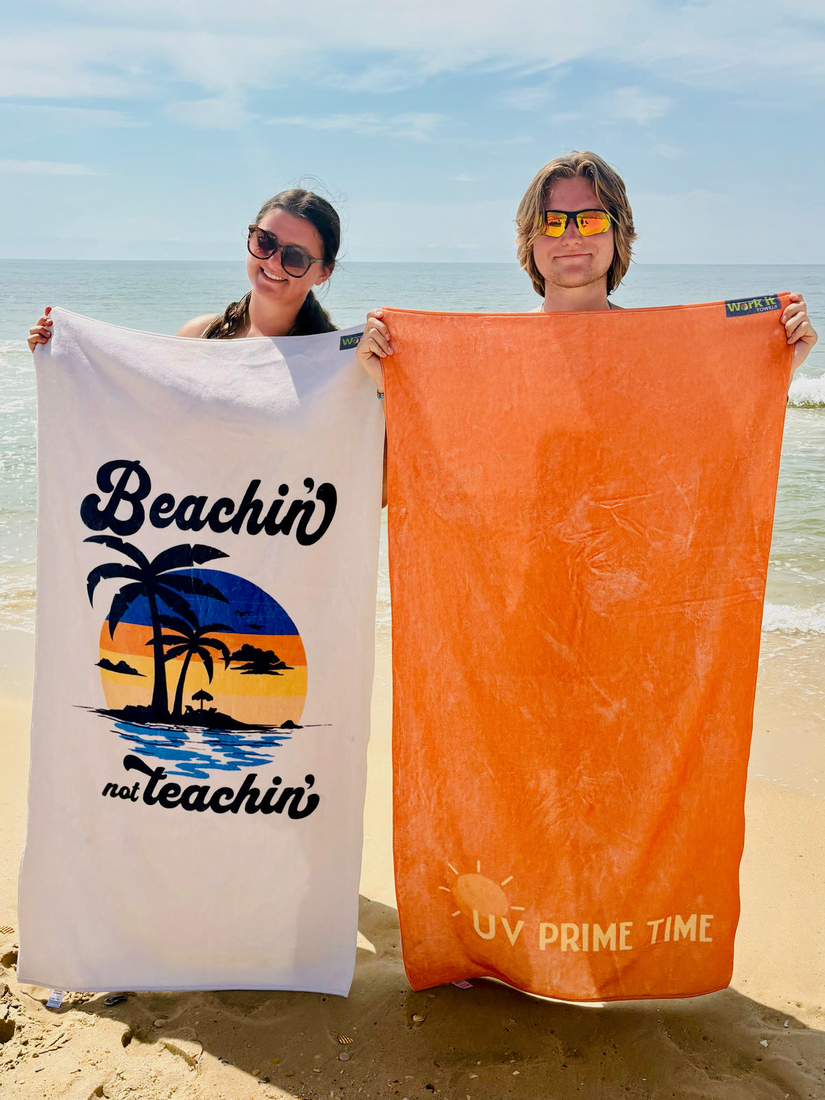 young woman holding the beachin not teachin towel with palm trees and young man holding the orange UV Prime Time beach towel design on a beach