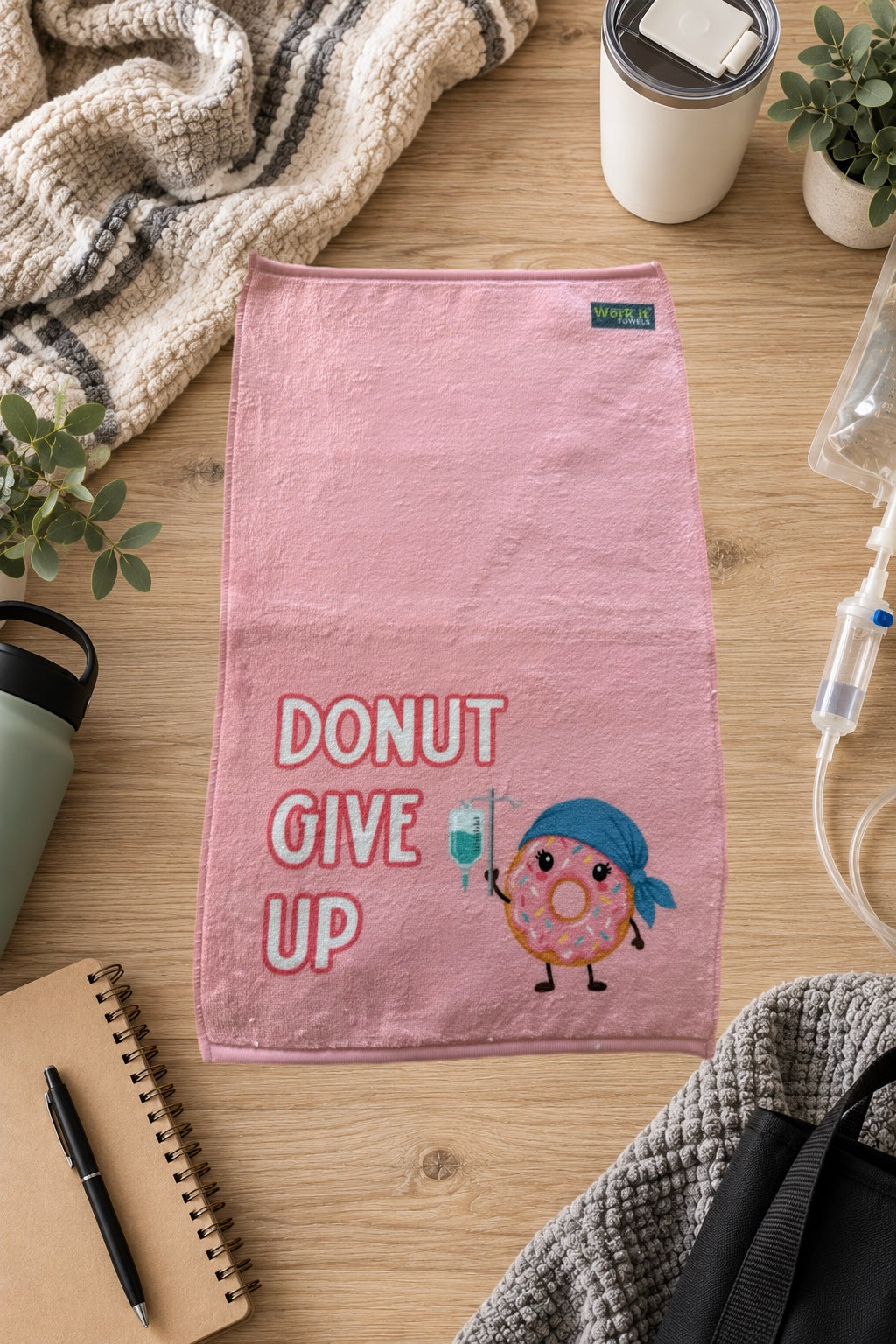plush pink towel with 'donut give up' and a pink iced donut wearing a cap and holding an IV on a hardwood floor surrounded by blankets, a notebook and drinks. 