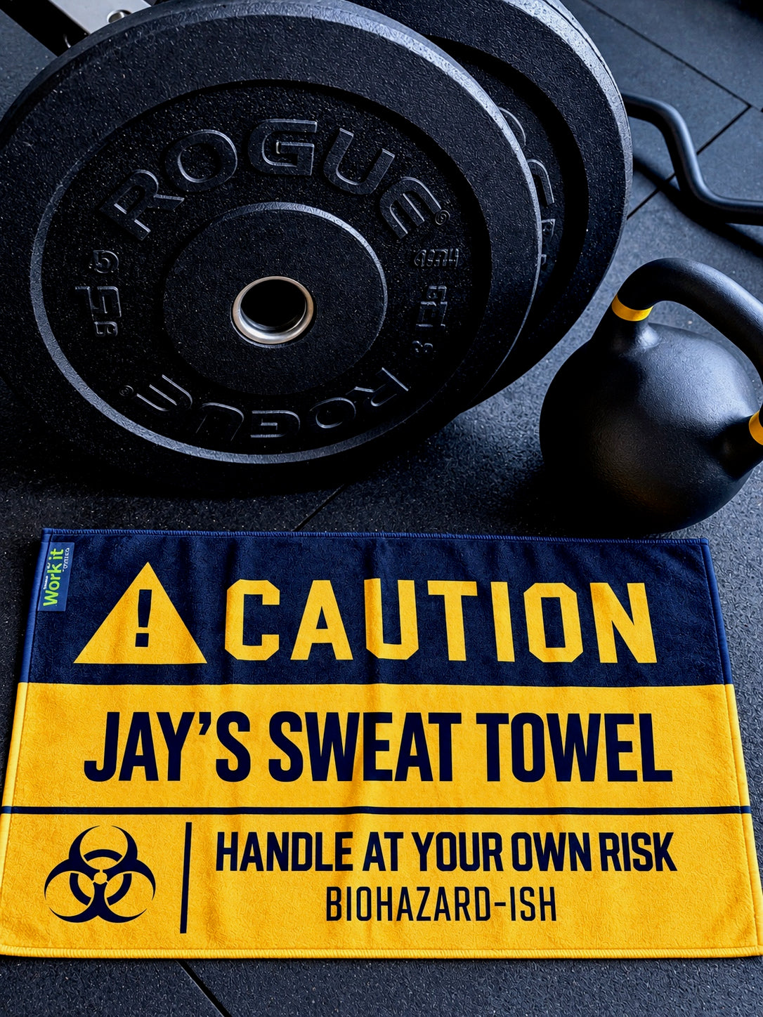 Personalized Work It Towels gym sweat towel with bold yellow and black CAUTION design, customized with “Jay's Sweat Towel” and “Handle at Your Own Risk” warning, displayed on a gym floor beside weight plates and a kettlebell.