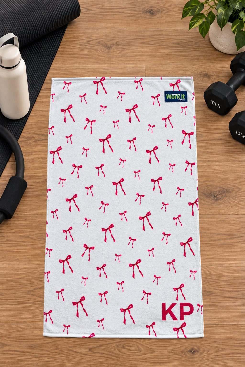 Pink bow personalized gym towel with custom 'KP' initials, laid flat on a wood floor next to fitness gear.