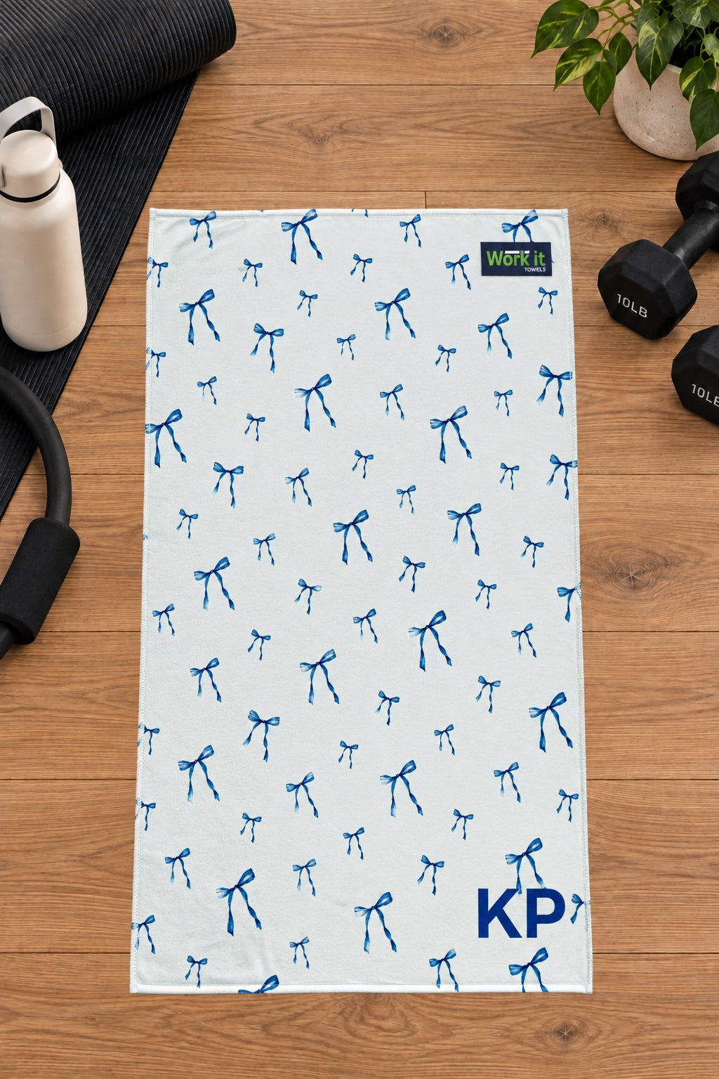 Blue bow personalized gym towel with custom 'KP' initials, laid flat on a wood floor next to fitness gear.