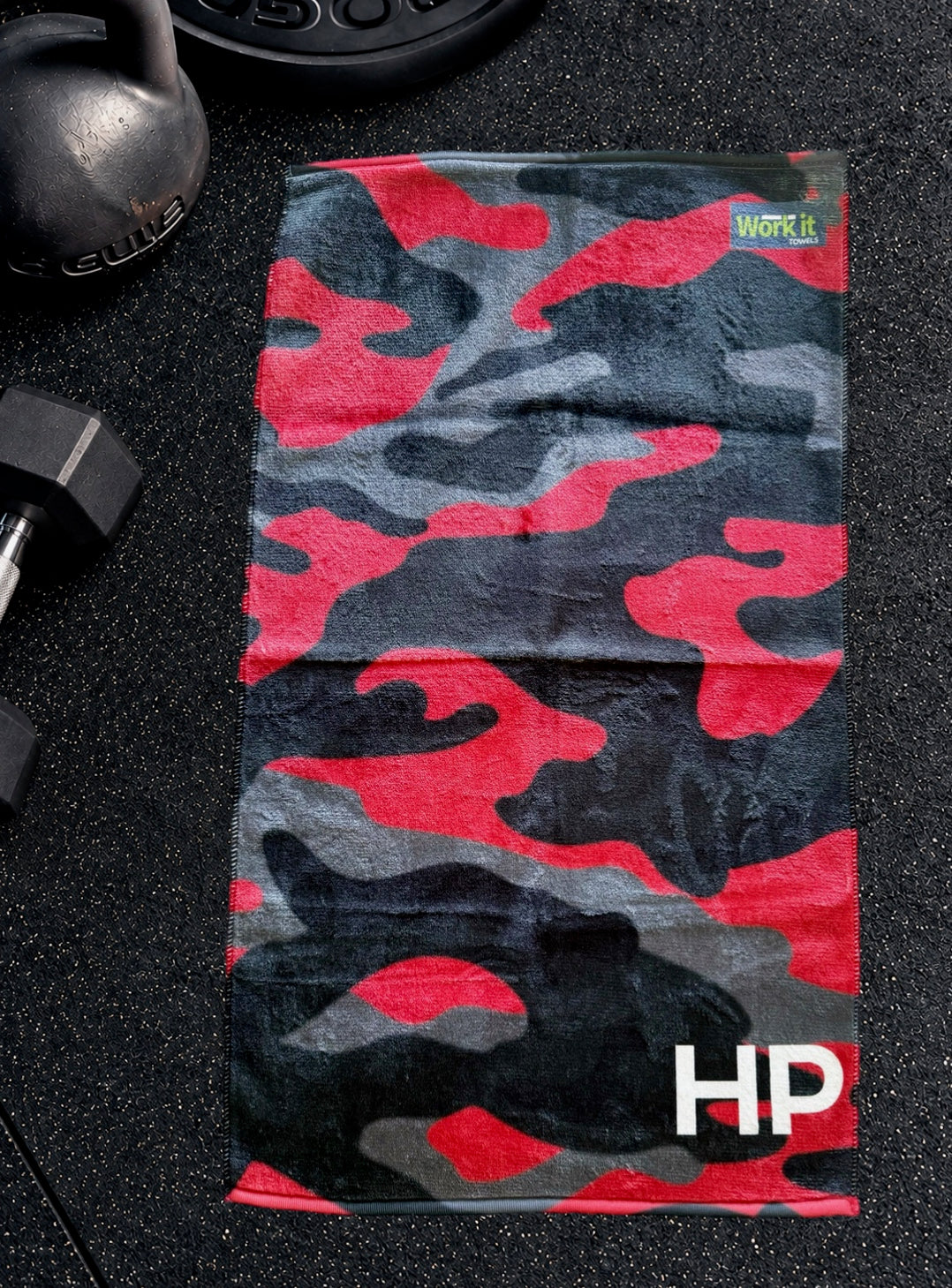Red camouflage personalized gym towel with custom 'HP' initials, laid flat on a black gym floor with a kettlebell and dumbbell.