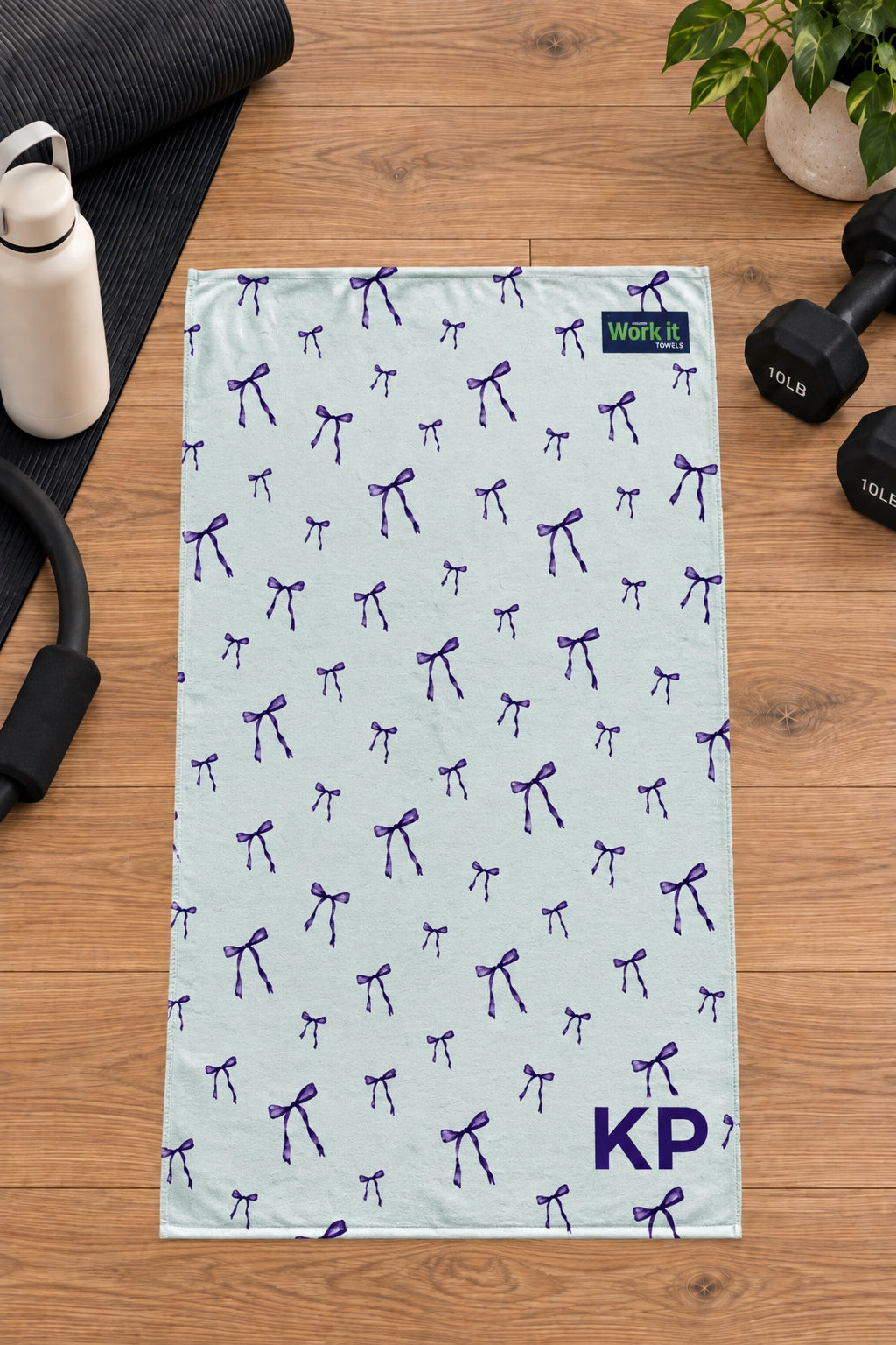 Purple bow personalized gym towel with custom 'KP' initials, laid flat on a wood floor next to fitness gear.