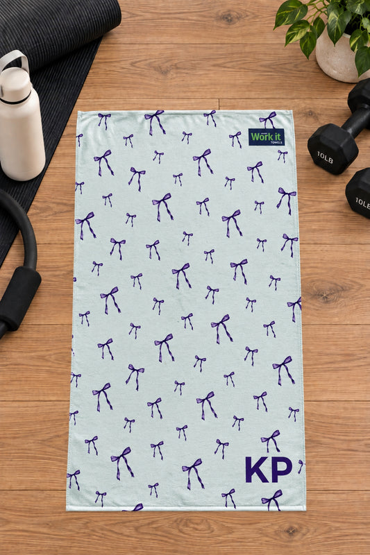 Purple bow personalized gym towel with custom 'KP' initials, laid flat on a wood floor next to fitness gear.