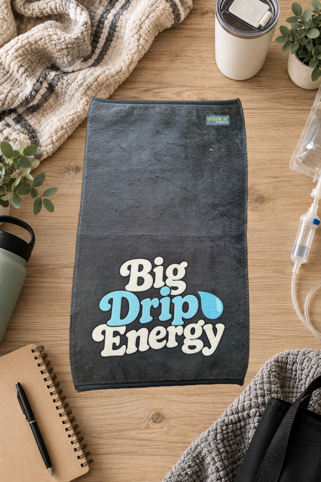 dark grey towel with 'big drip energy' text and a tear drop on a hardwood floor with blankets and drinks