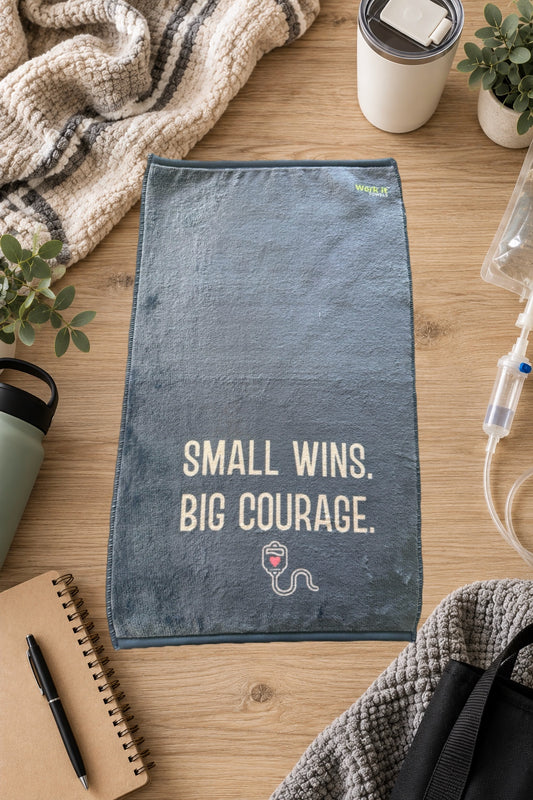 blueish grey plush towel with text, 'small wins, big courage,' and a small IV graphic with a heart on a hardwood floor with blankets and drinks