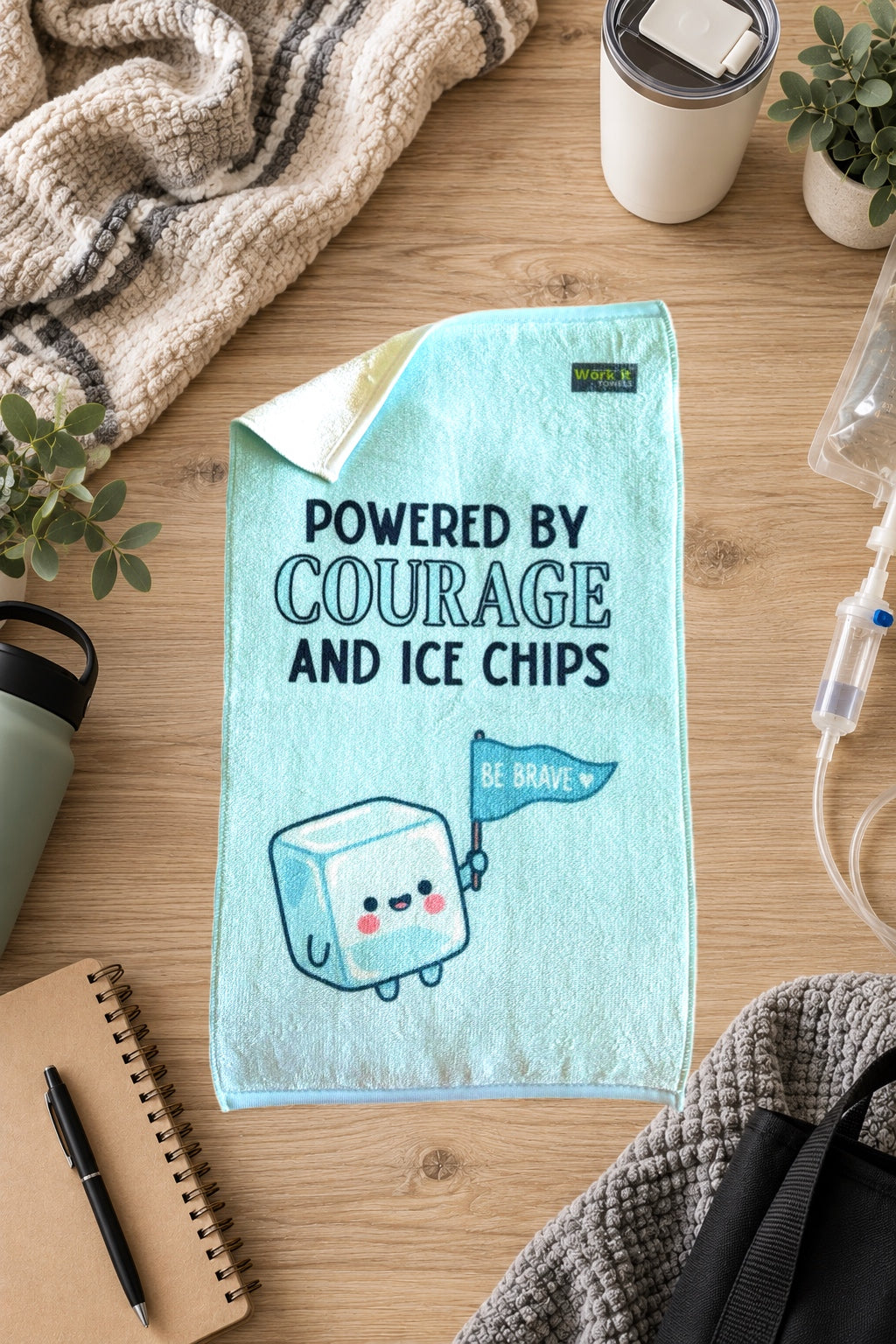 A light blue towel laid flat on a wooden surface, printed with the phrase 'POWERED BY COURAGE AND ICE CHIPS' and a cute ice cube character holding a 'BE BRAVE' flag. The scene is styled with a blanket, plants, a tumbler, notebook, pen, and other everyday props.