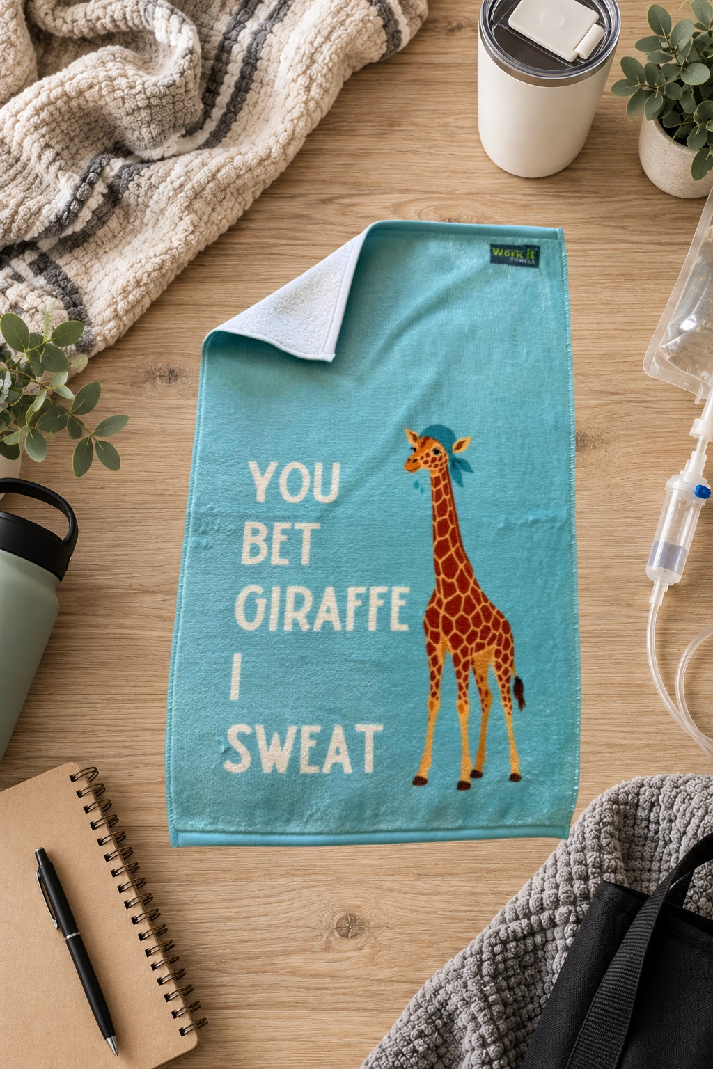 a light blue towel with a giraffe wearing a cap and sweating with text, 'you bet giraffe I sweat,' on a hardwood floor with blankets and drinks. 