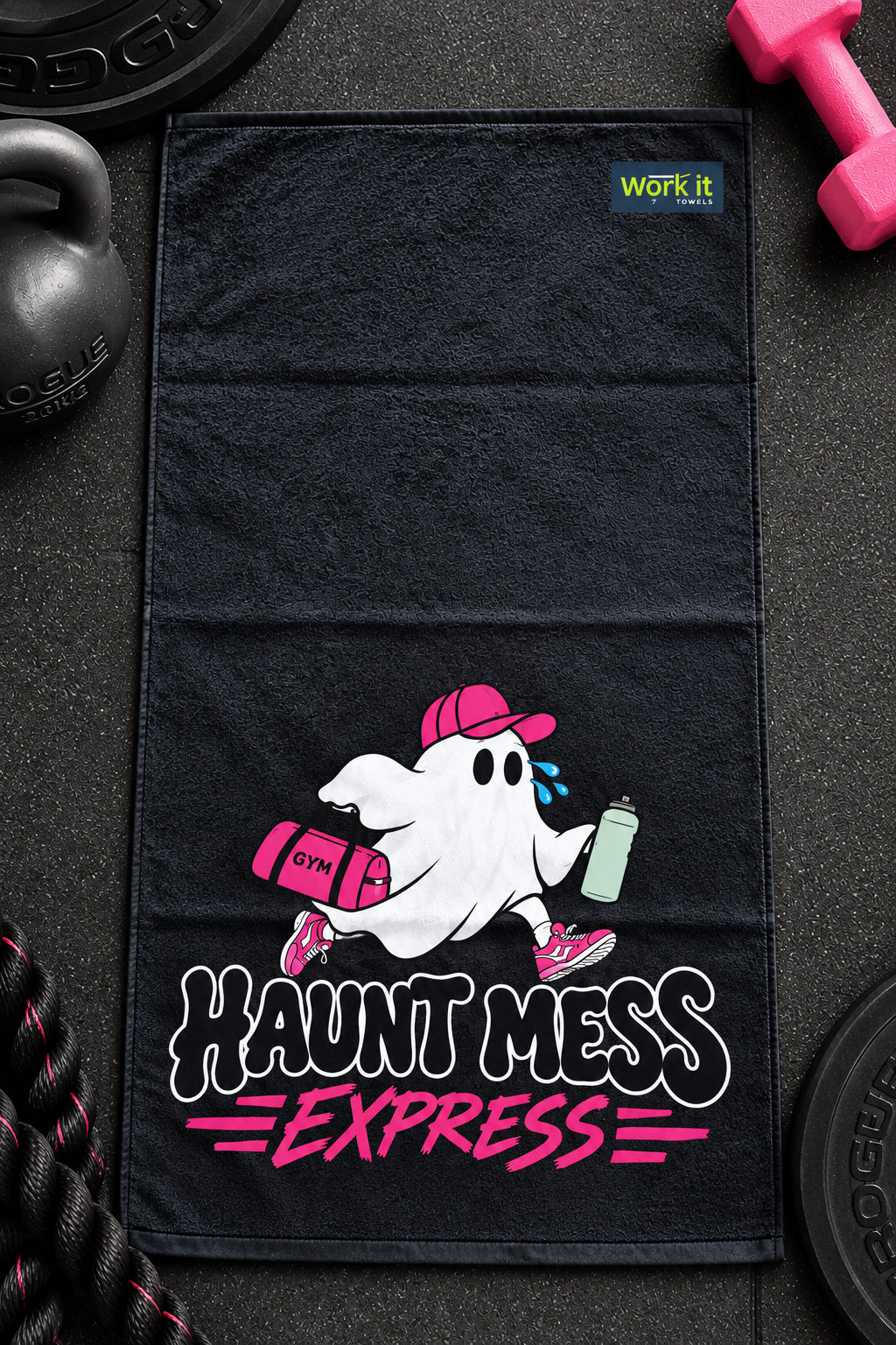 A black gym towel laid flat on a dark textured surface, featuring a cartoon ghost wearing a pink cap and carrying a pink bag and water bottle, with the text 'HAUNT MESS EXPRESS' in white and pink. A small 'WORK IT' logo is printed near the top right corner of the towel.