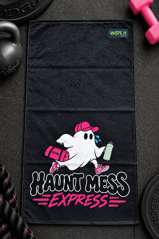 A black gym towel laid flat on a dark textured surface, featuring a cartoon ghost wearing a pink cap and carrying a pink bag and water bottle, with the text 'HAUNT MESS EXPRESS' in white and pink. A small 'WORK IT' logo is printed near the top right corner of the towel.