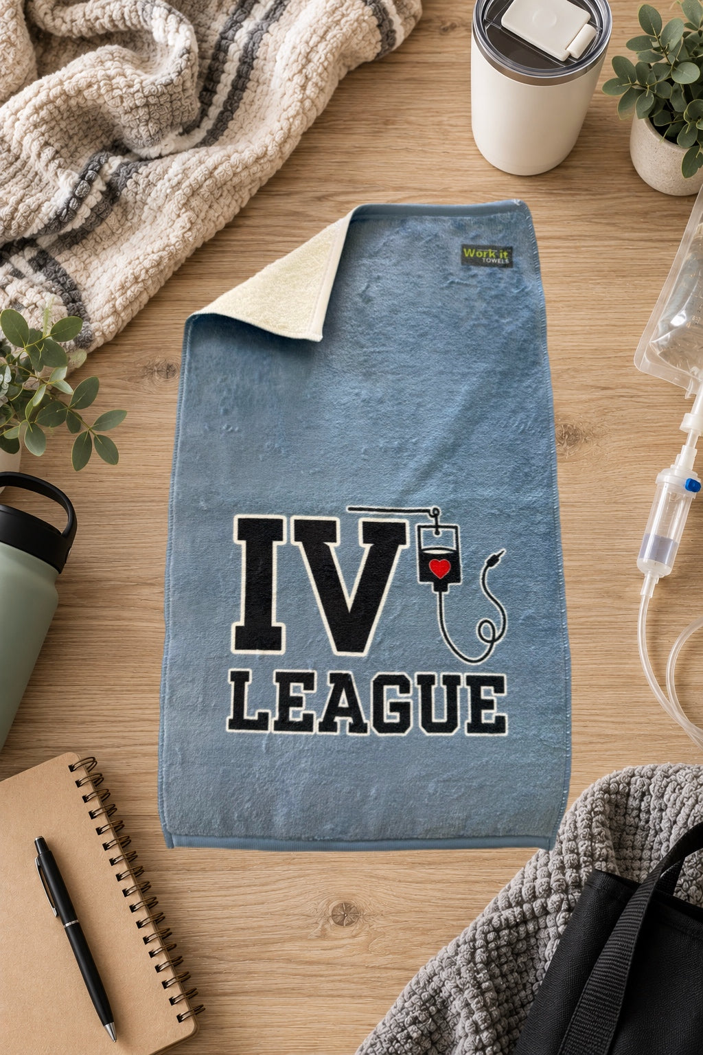 blueish grey towel with text, 'IV League' and an IV hanging from the V.  The towel is laying on a hardwood floor with blankets and IVs