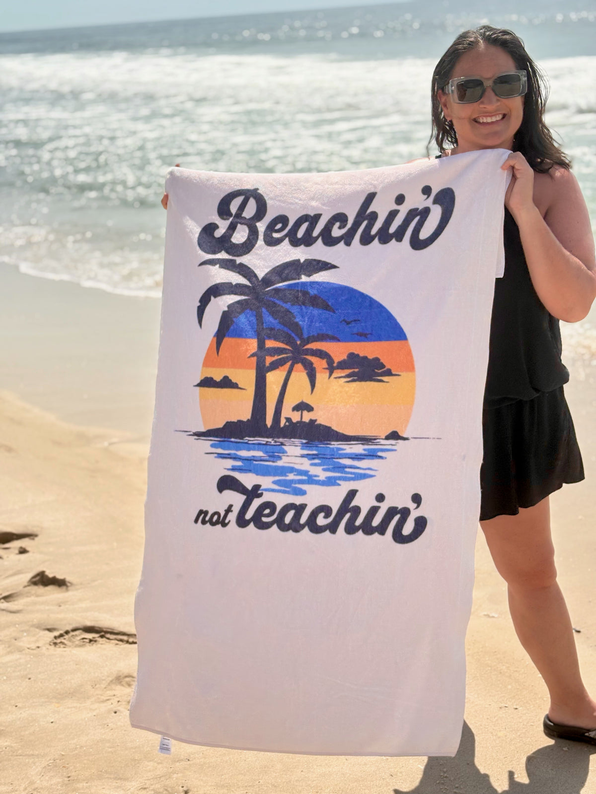 our 'beachin not teachin' sunset design beach towel being held by a young woman on a beach 