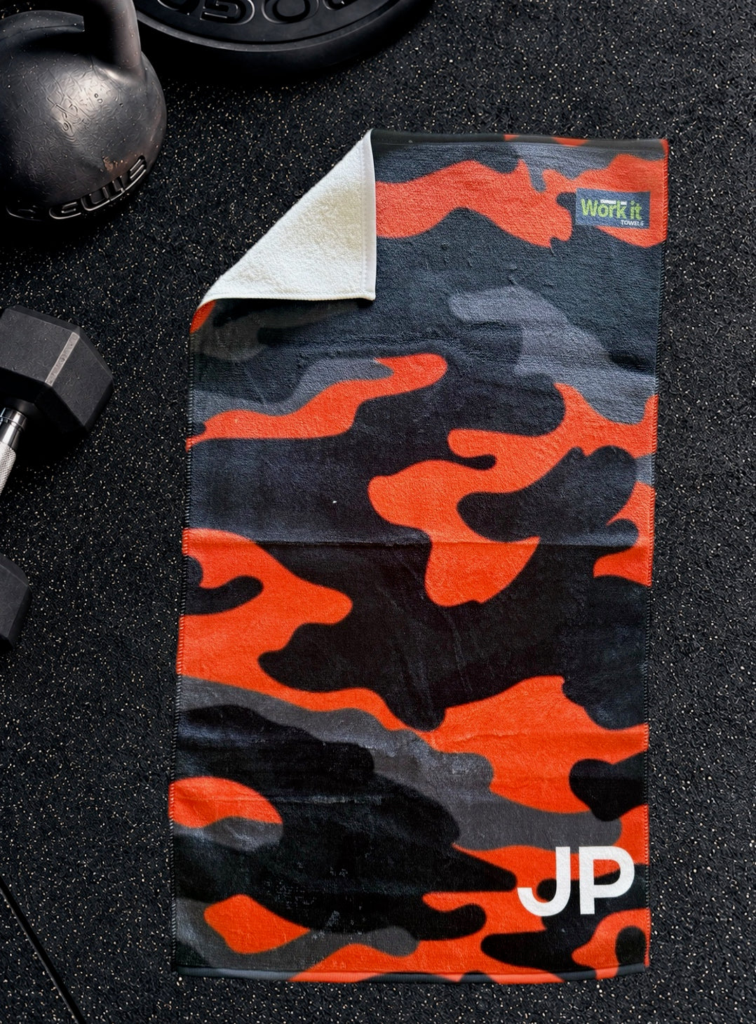 Orange camouflage personalized gym towel with custom 'JP' initials, laid flat on a black gym floor with weights.