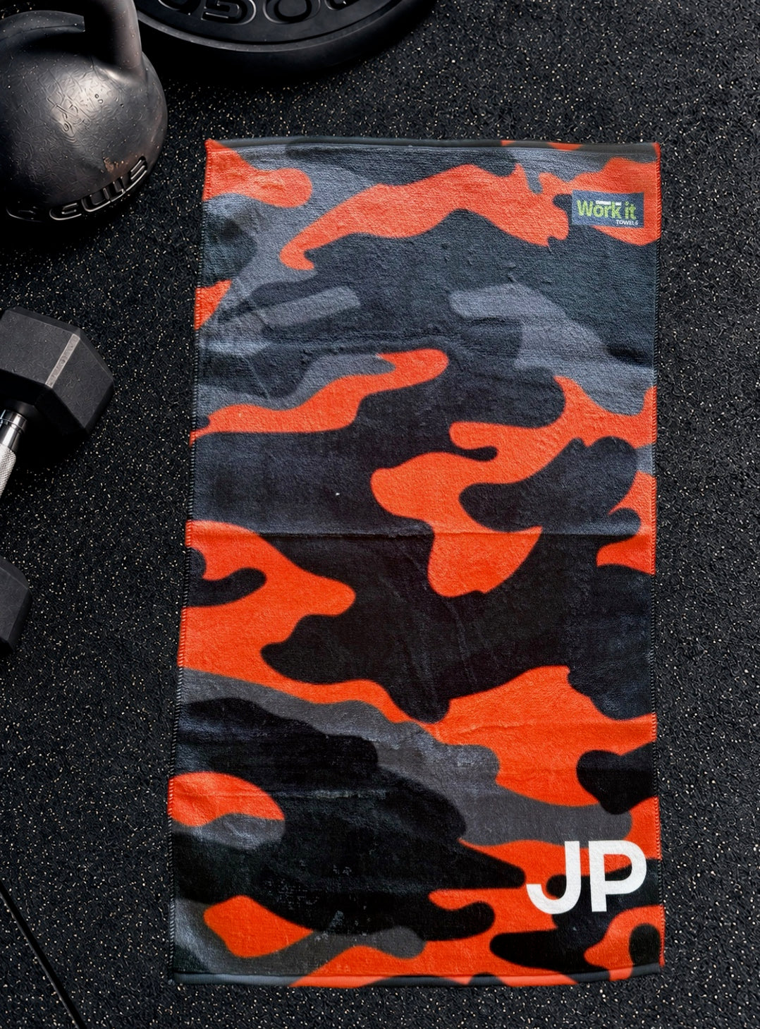 Orange camouflage personalized gym towel with custom 'JP' initials, laid flat on a black gym floor with a kettlebell and dumbbell.